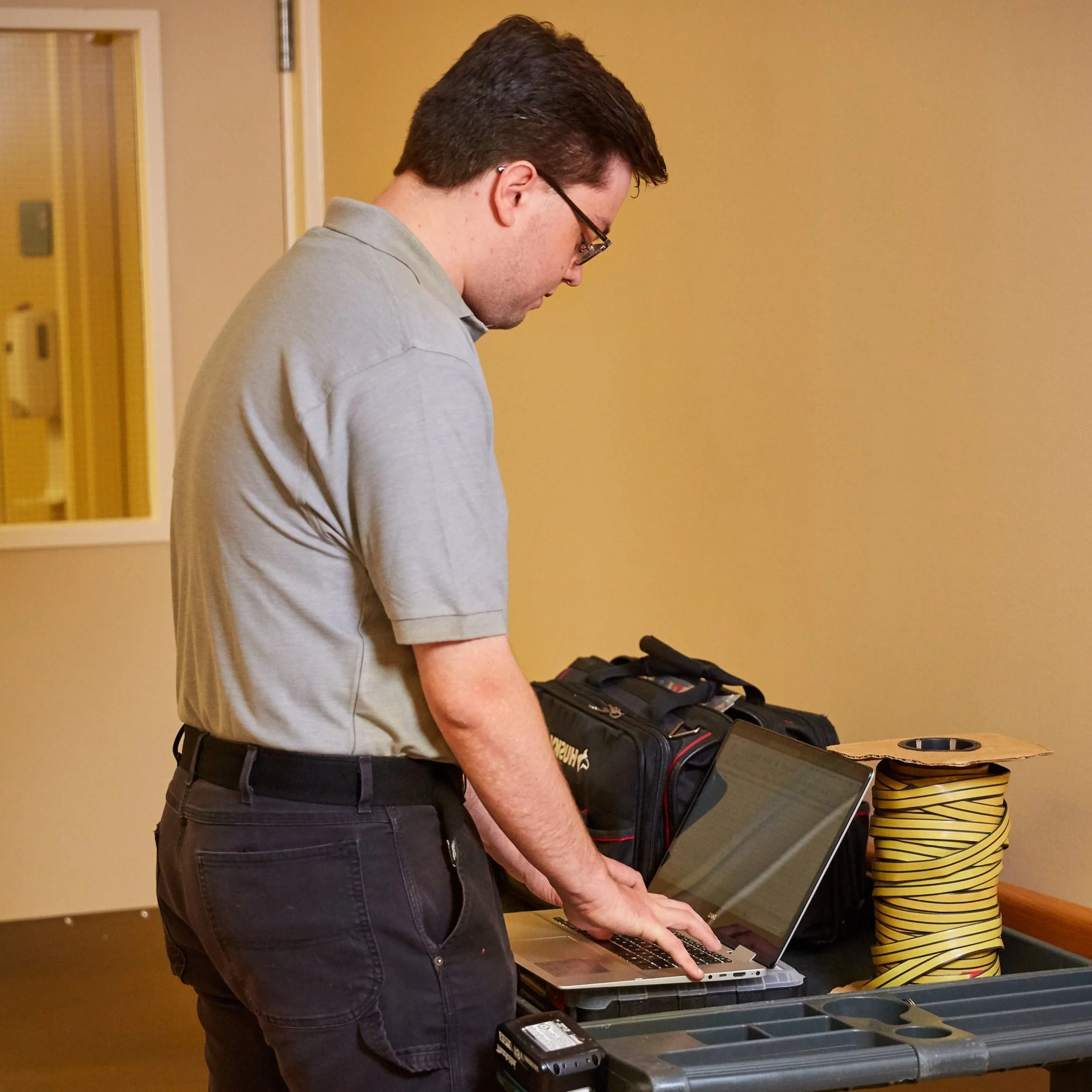 A man wearing a gray polo shirt and black pants working on a laptop at a table with equipment and supplies.
