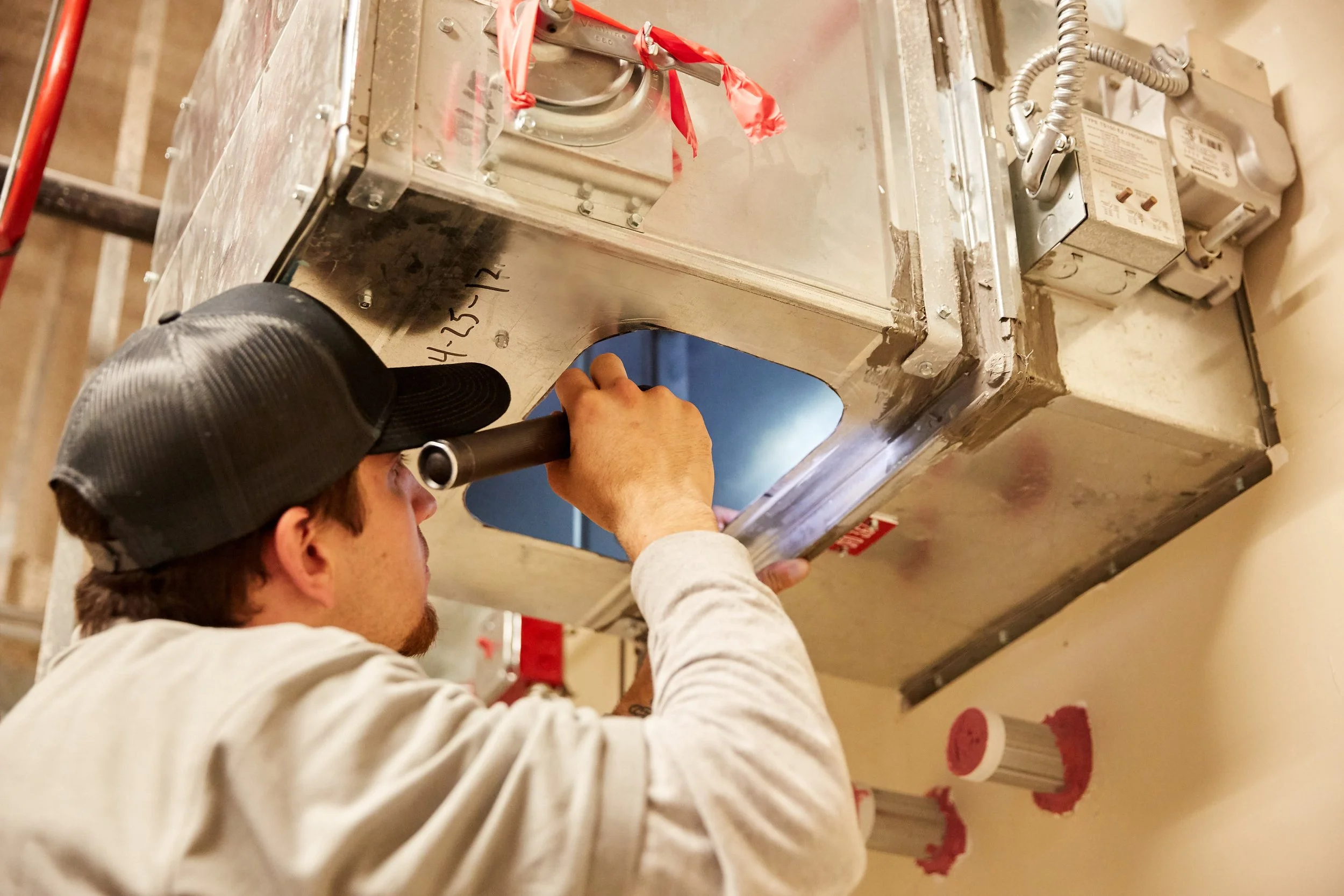 A man performing inspection on a damper , using a flashlight, in a hospital setting.