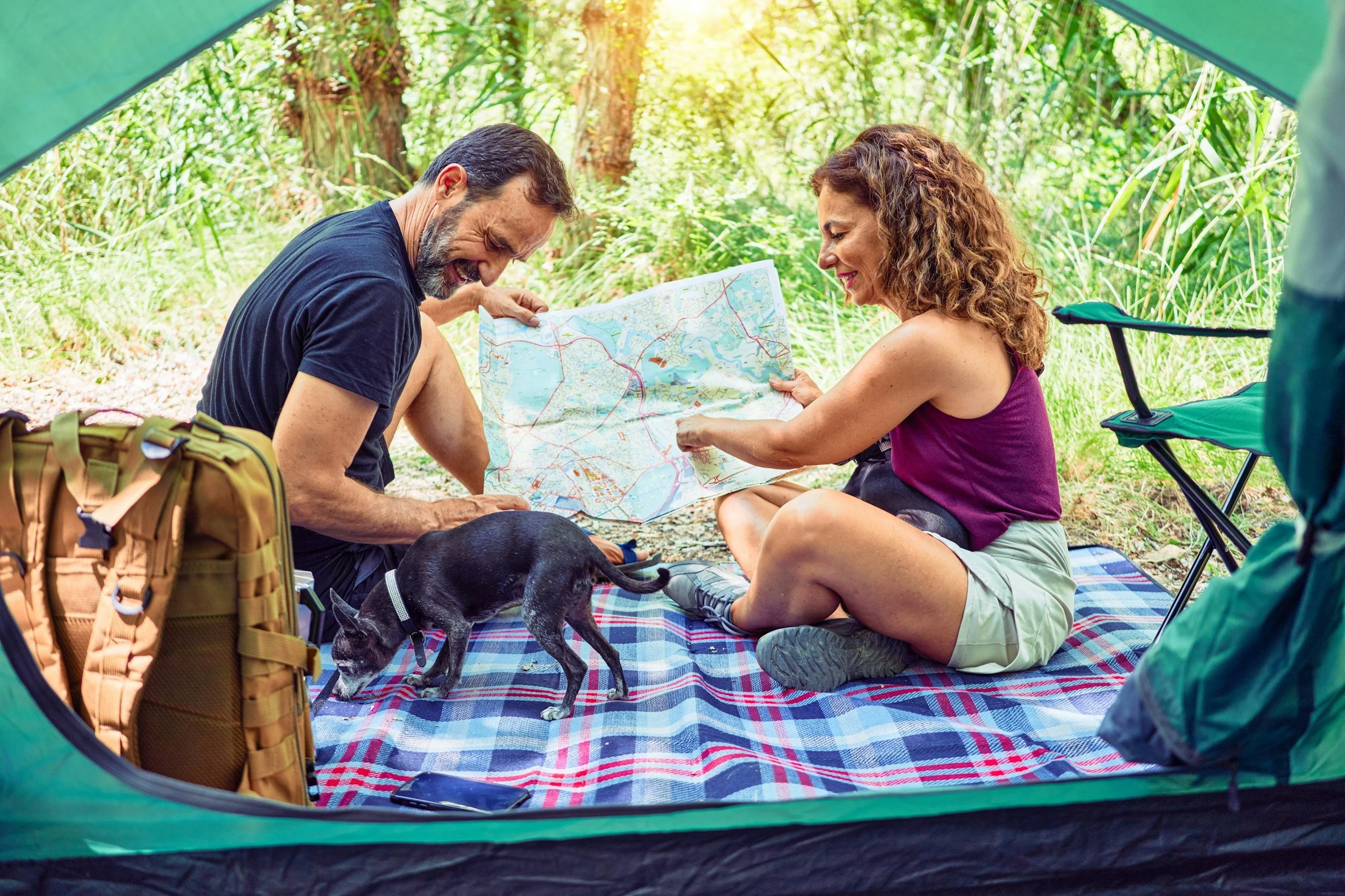 Two people, a man and a woman, sitting on a blanket inside a camping tent in a forest. They are looking at a map and laughing. A small black dog with a collar is sniffing the blanket, and a backpack is nearby.