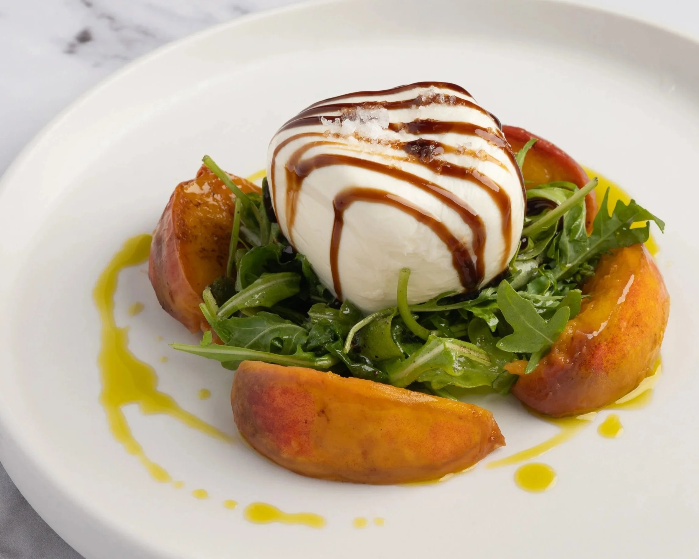 Salad with arugula, grilled peaches, burrata cheese topped with balsamic glaze, garnished with olive oil and sea salt on a white plate.