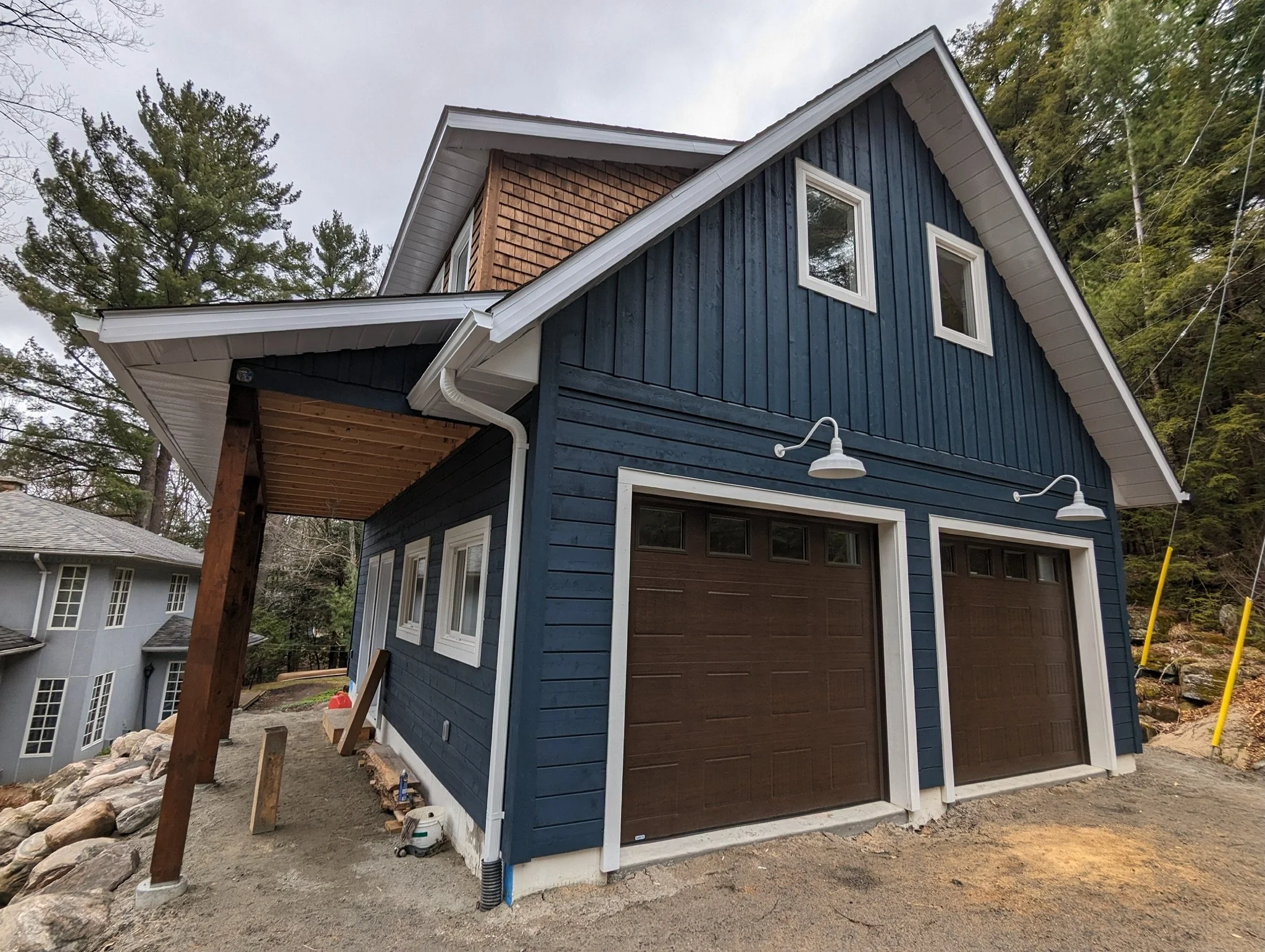 A blue two-story house with white trim and two brown garage doors. It features a sloped roof, exterior lighting fixtures above the garages, and is surrounded by trees and rocky landscaping.