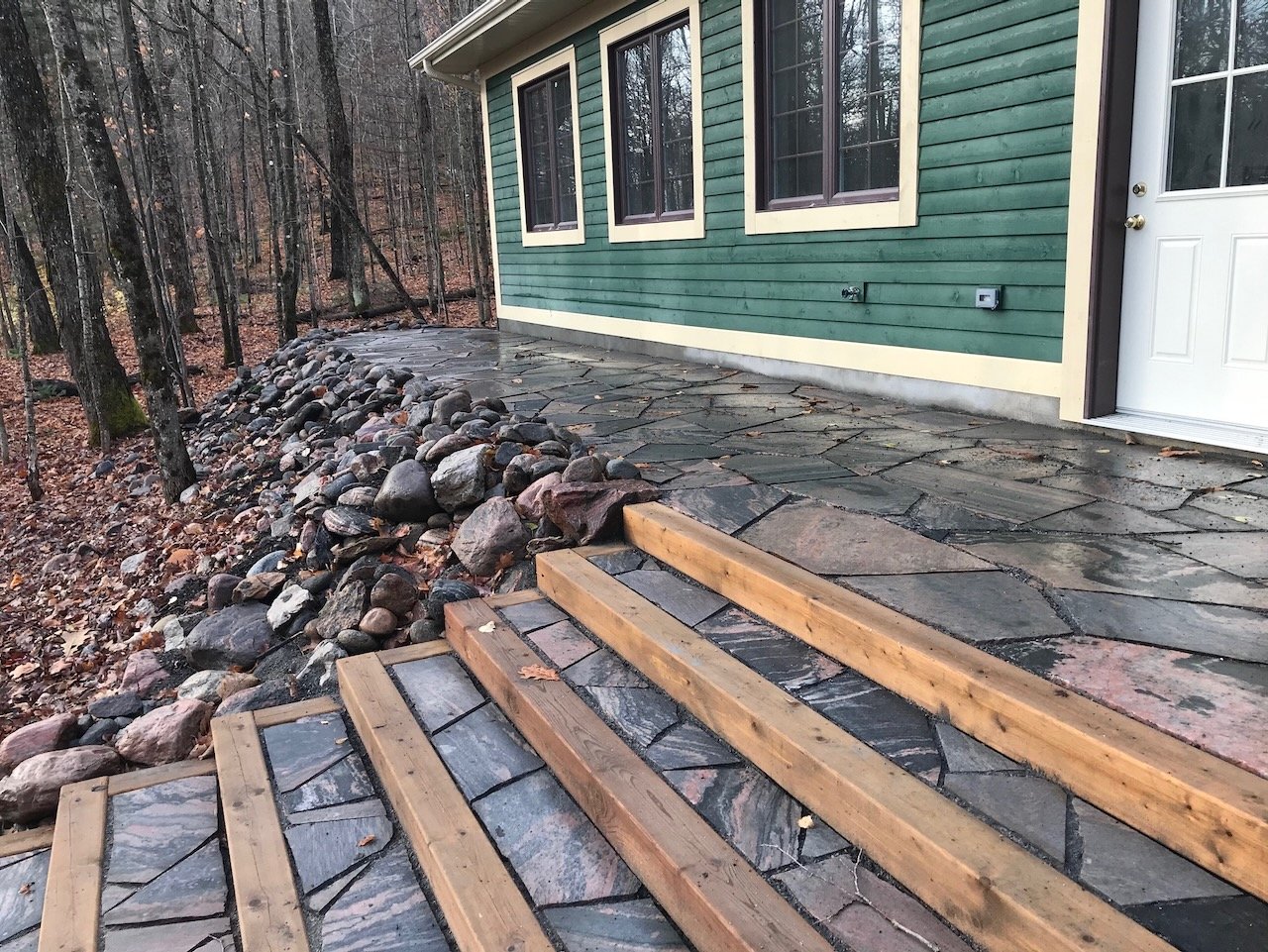 Stone patio and steps next to a green house in a forested area, with a rock border.