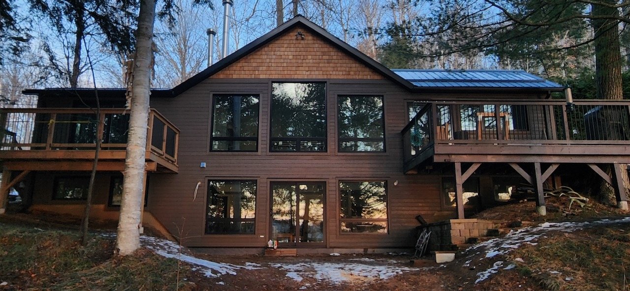 A two-story wooden cabin with large windows, surrounded by trees and minimal snow on the ground, featuring a deck on the right side.
