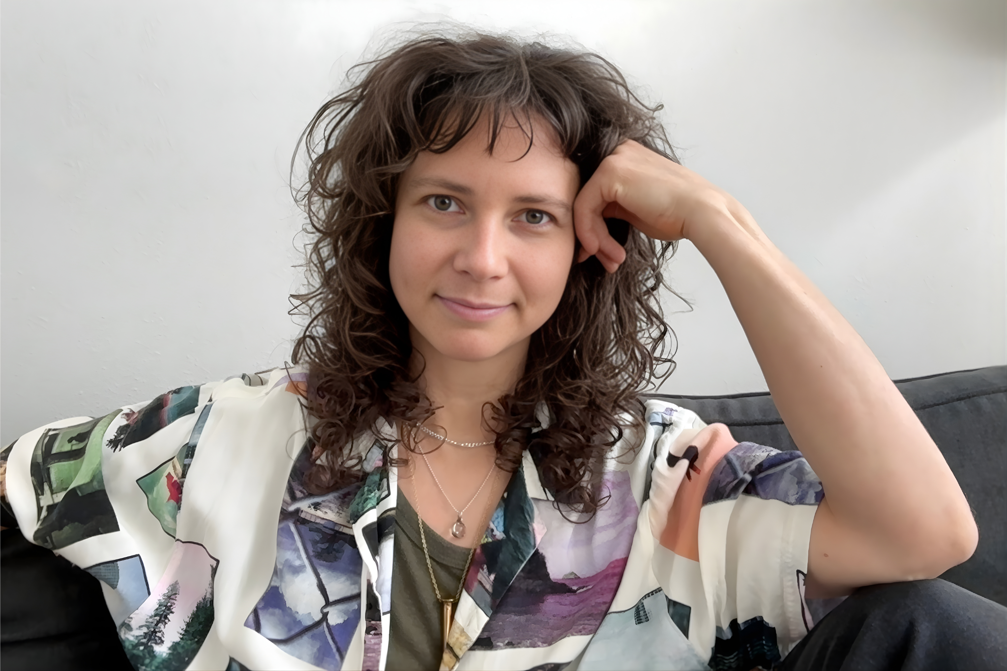 A therapist with brown, wavy hair sitting on a couch with a relaxed pose, smiling gently, wearing a colorful patterned shirt, a necklace, and a dark green top underneath, in a room with a white wall background.