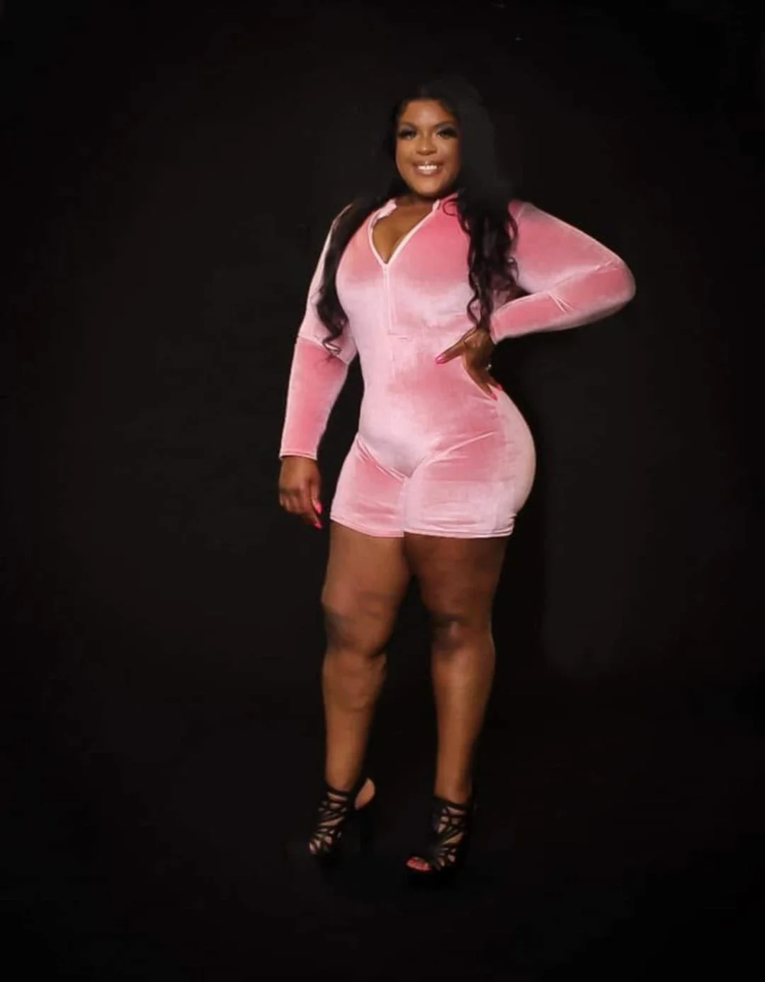 A woman with long dark hair in a pink velvet jumpsuit and black lace-up high heels standing against a black background.