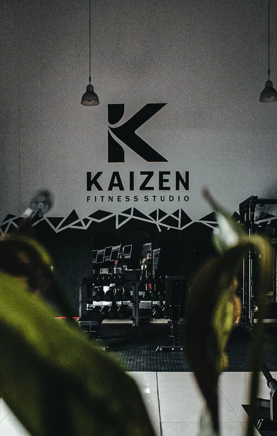 Interior of Kaizen Fitness Studio with gym equipment visible, wall logo featuring a large black letter 'K' and smaller text 'KAIZEN FITNESS STUDIO' beneath, surrounded by black geometric designs.