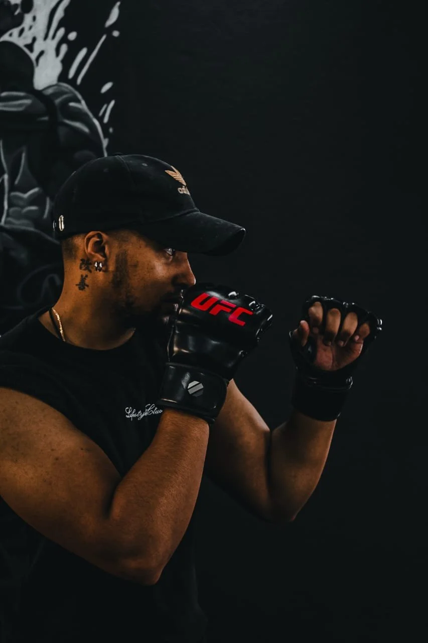 A male UFC fighter in a fighting stance, wearing black gloves with red UFC logo, a black cap, and sleeveless shirt, standing against a dark background.