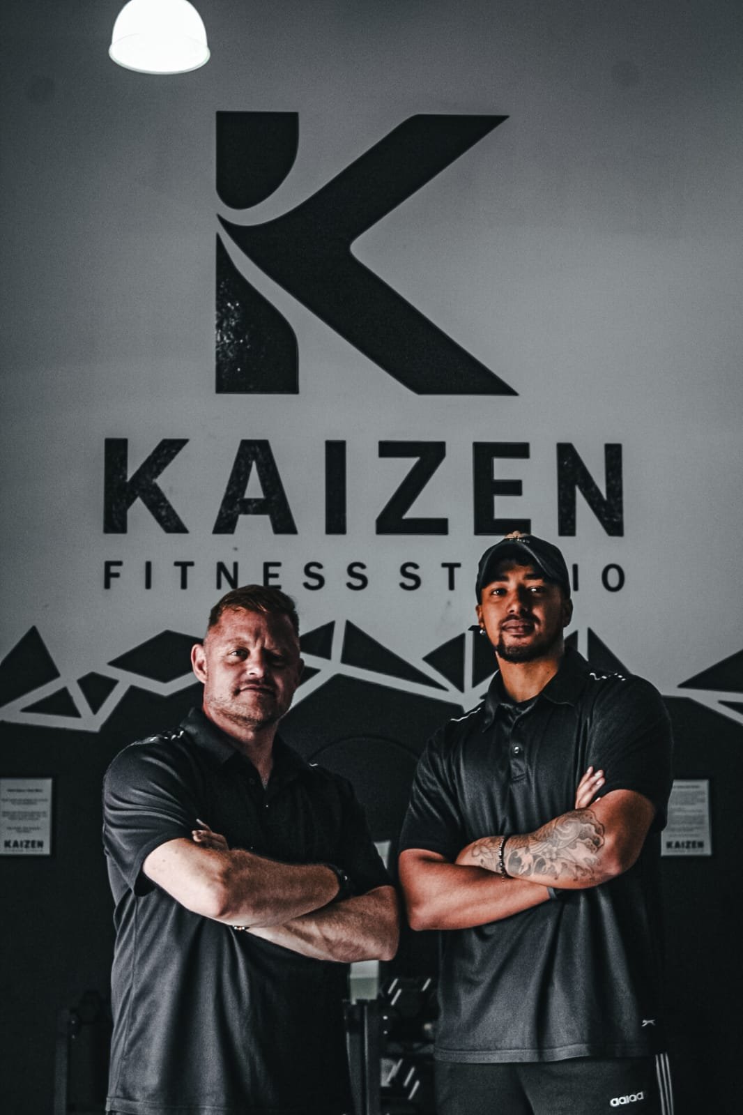 Two men standing with arms crossed in front of a wall with the logo and text 'KAIZEN FITNESS STUDIO,' inside a gym or fitness studio.