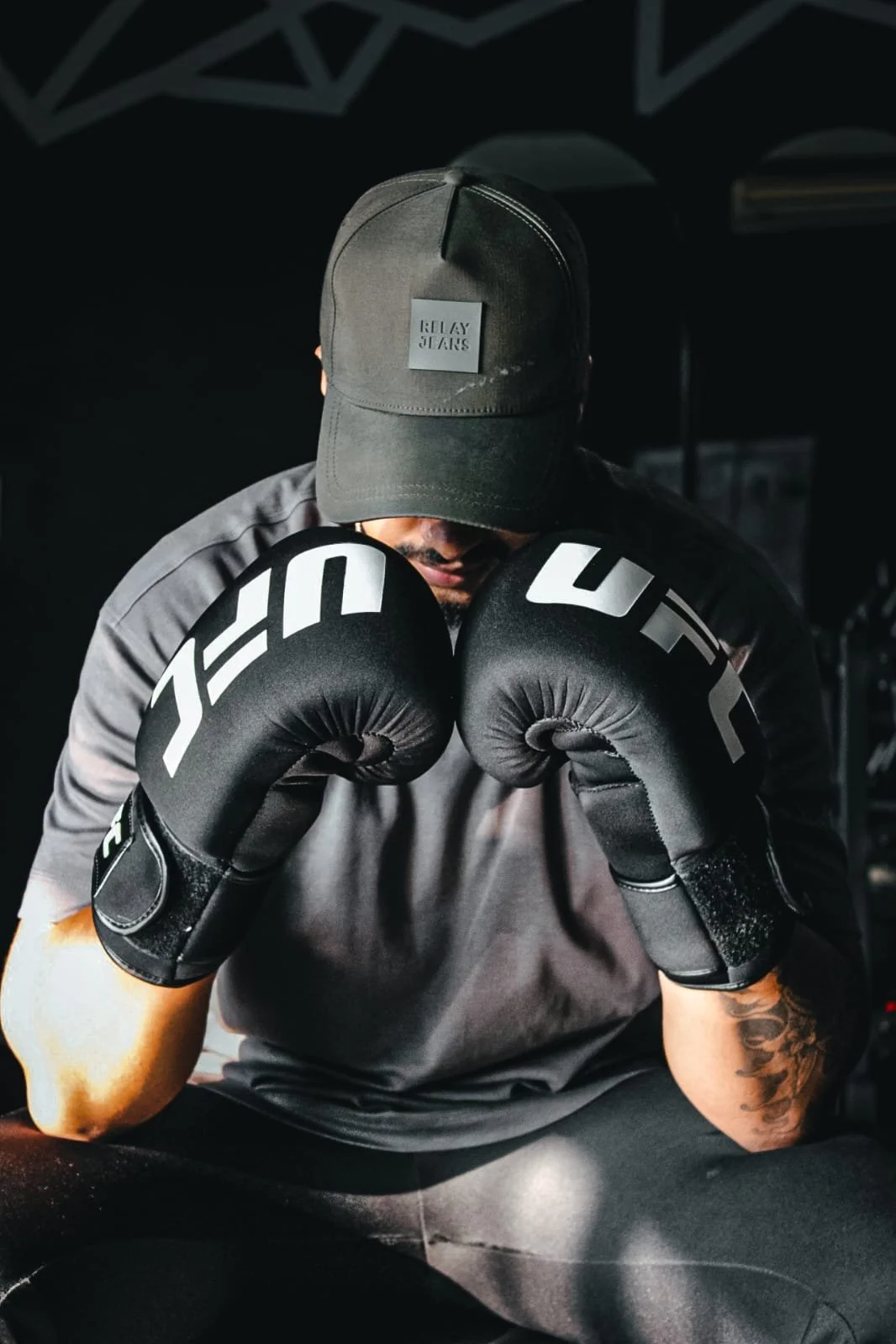 A boxer wearing black Focus Mitts with the word "UFC" printed on them, a gray t-shirt, and a black cap with the words "Relay Stars" embossed, is in a boxing stance with their head down, in a dark gym environment.