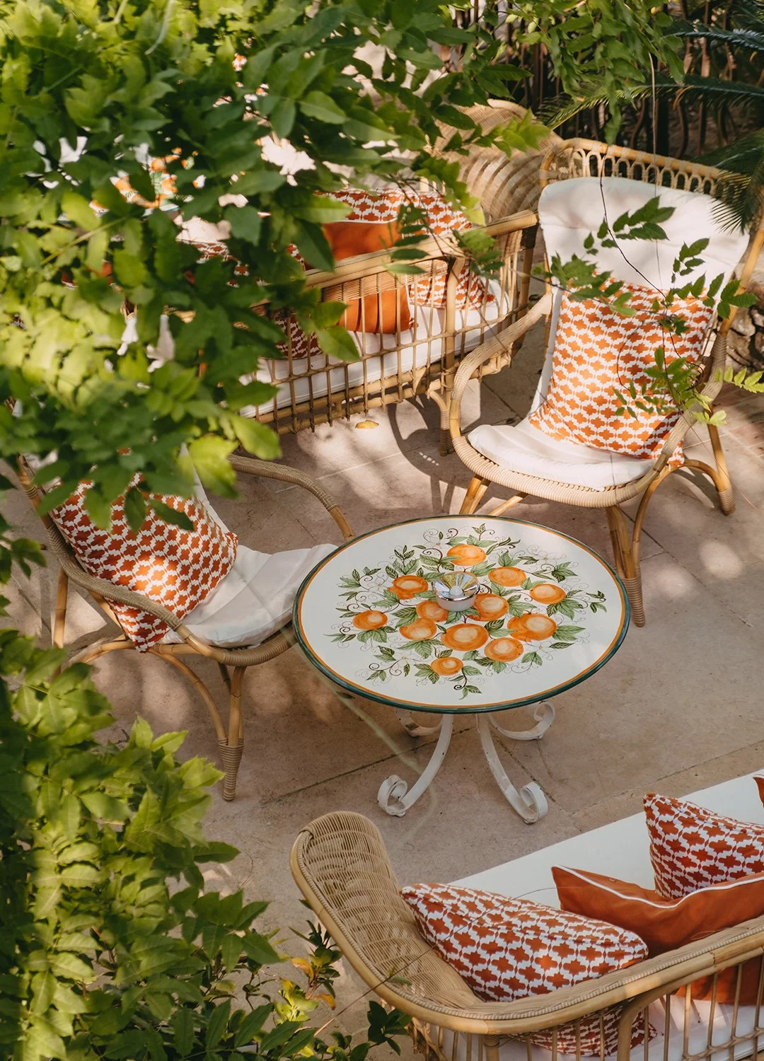 Outdoor patio furniture set with rattan chairs, a glass-topped table with orange fruit design, surrounded by greenery and cushions in orange patterned fabric.