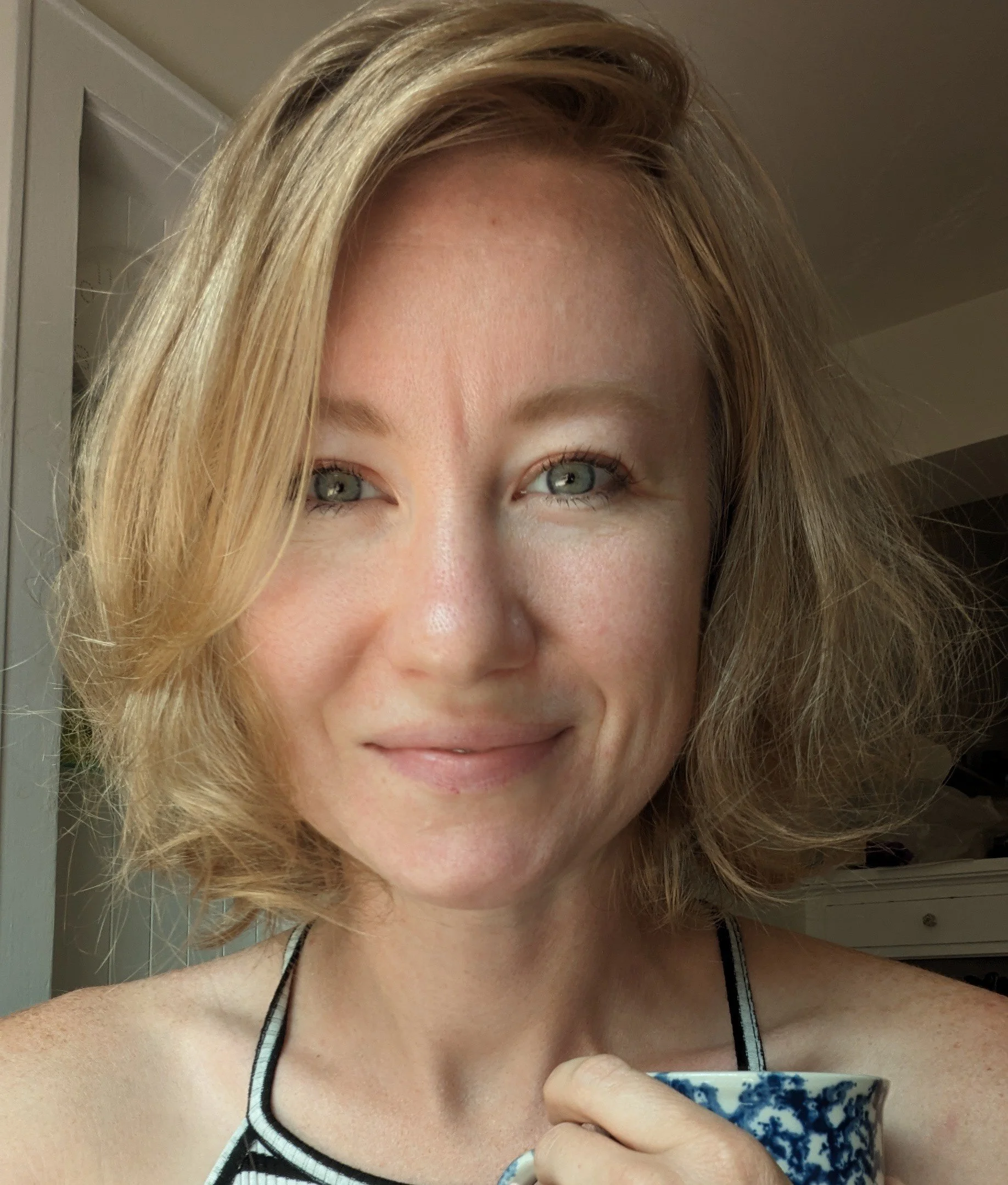 Close-up of a woman with blonde hair, blue eyes, and light skin in a room with cream walls. She is smiling slightly and wearing a black and white striped tank top with a blue and white patterned cup in her hand.