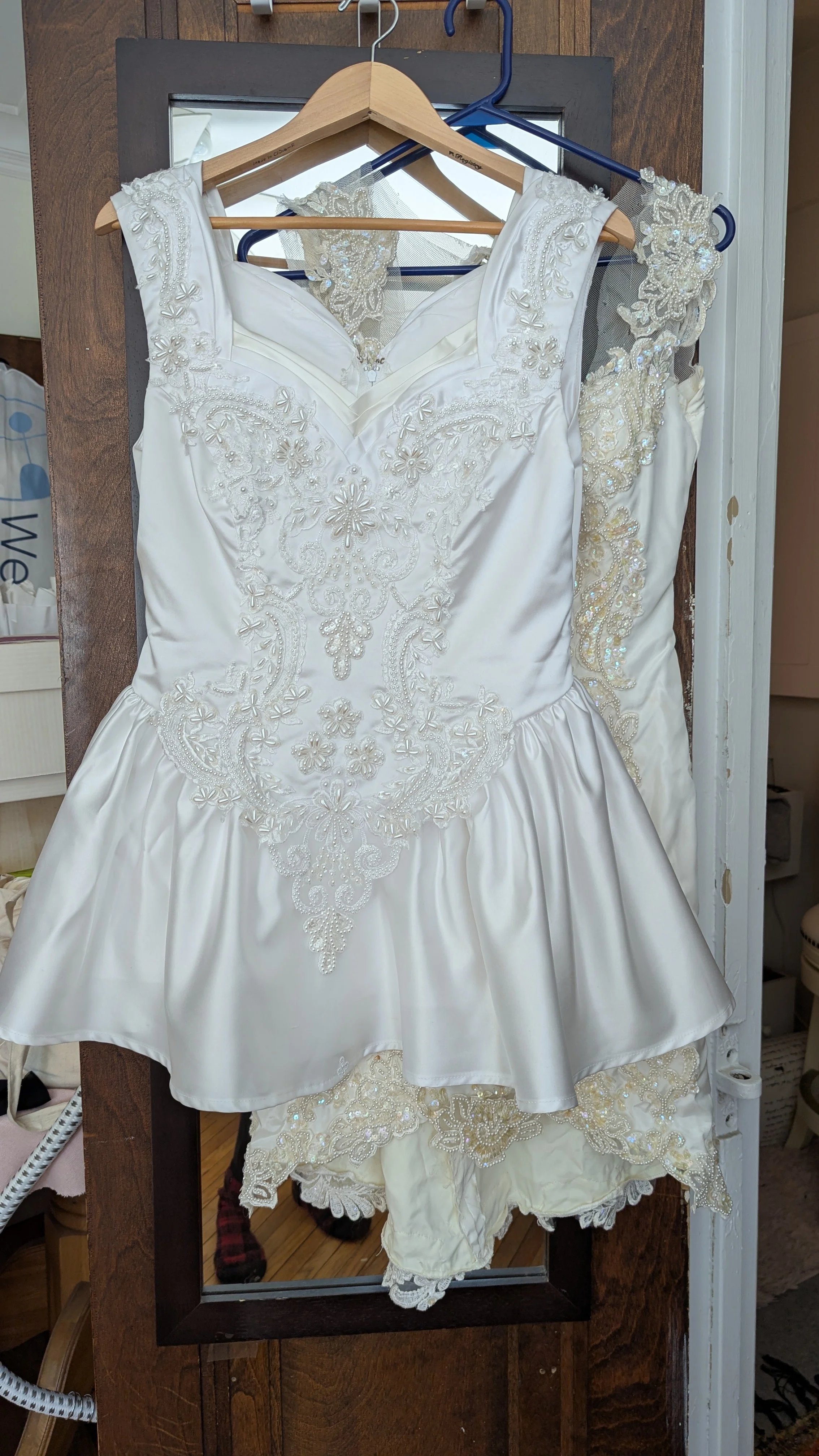 A wedding dress with intricate embroidery hangs on a hanger, with lace and beaded detailing on the shoulder and bodice, a separate lace veil in the background, and matching lace shorts hanging below.