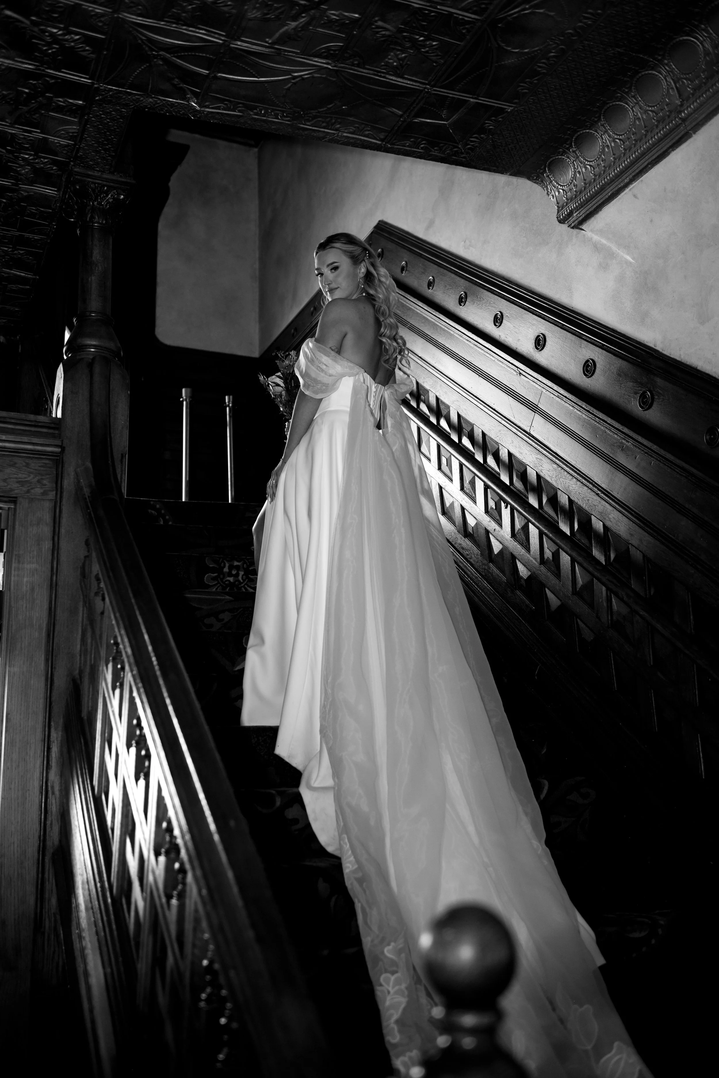 A woman in a wedding dress standing on a staircase inside a historic building with ornate woodwork and decorative ceiling details, looking back over her shoulder.