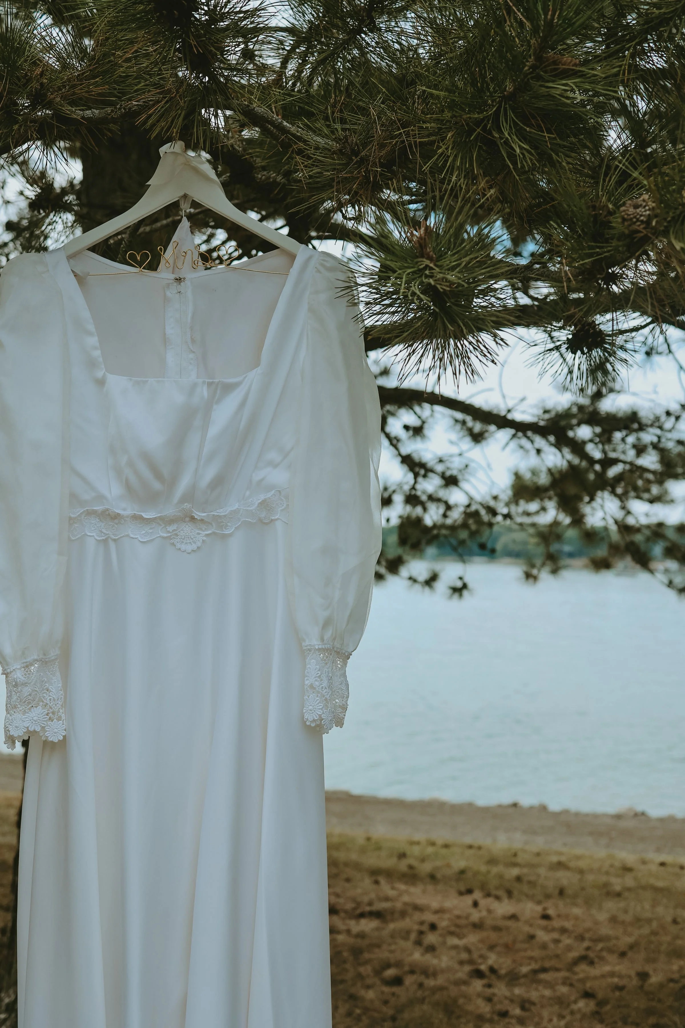 A white wedding dress hangs on a hanger with a gold heart-shaped hanger in the shape of 'Mrs.' attached to the collar, hanging from a pine tree branch. In the background, there is a lake and a sandy shoreline.