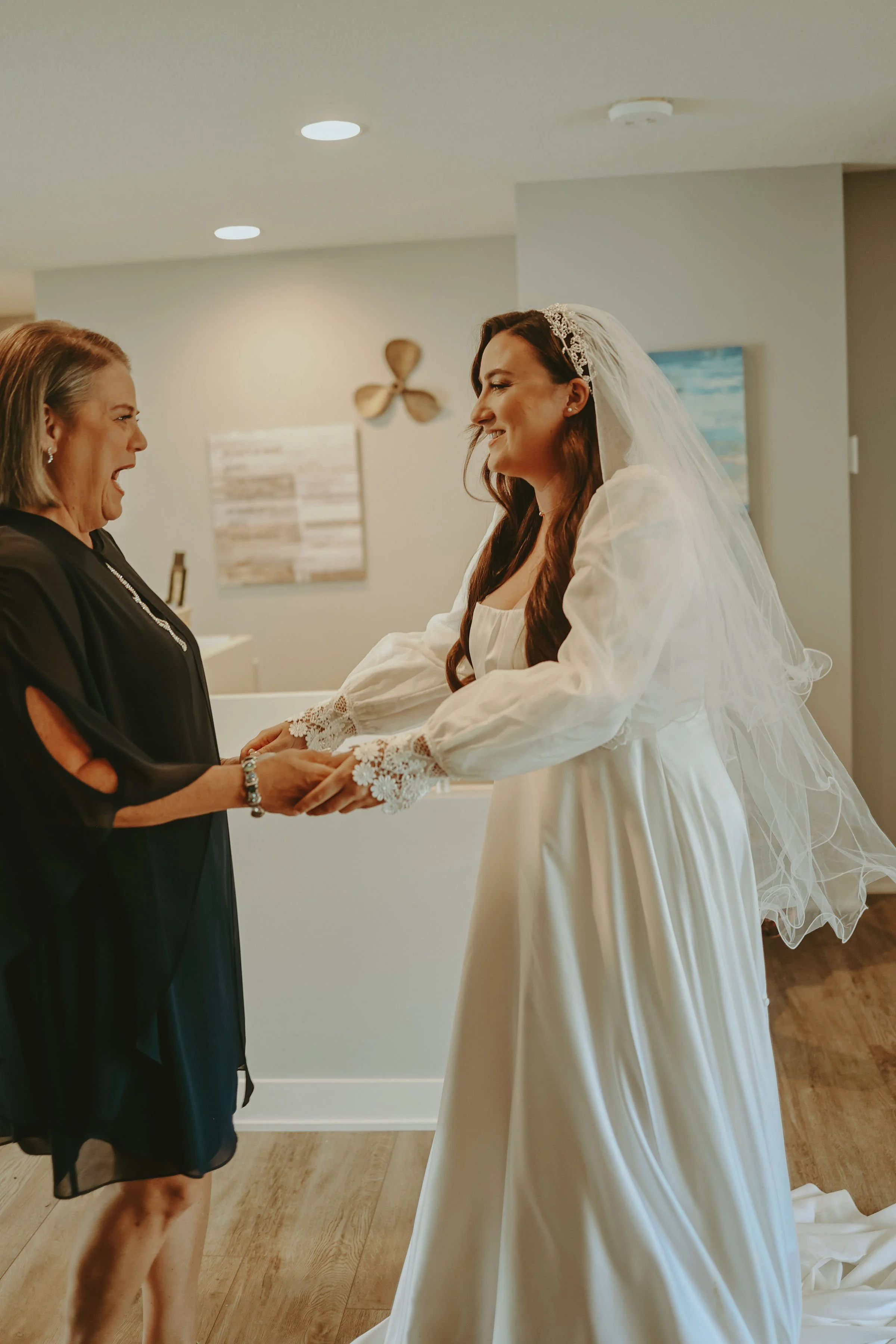A bride in a white wedding dress with lace sleeves and a veil holding hands with her mother in a black dress, both smiling, in a room with a wooden floor, painting, and wall decor.