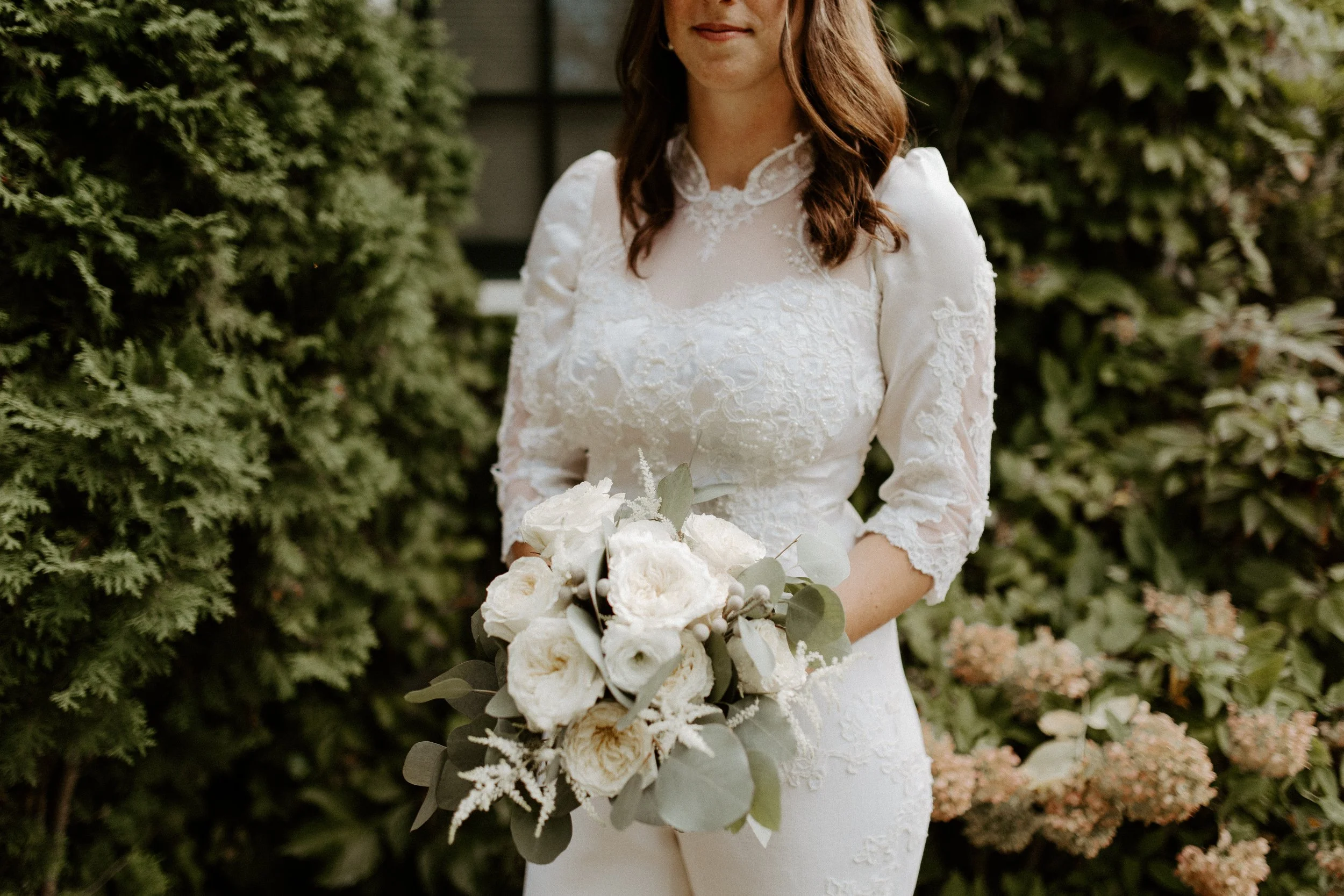 A woman in a white lace wedding dress holding a bouquet of white roses and greenery outdoors with a background of green bushes.