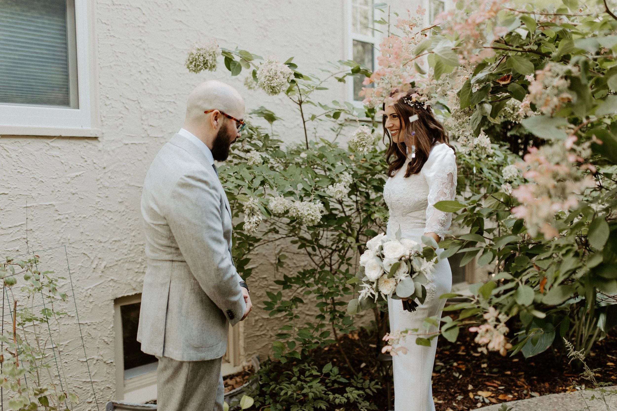 A wedding ceremony with a bride and groom standing outside among pink and white flowers, holding hands and smiling at each other.