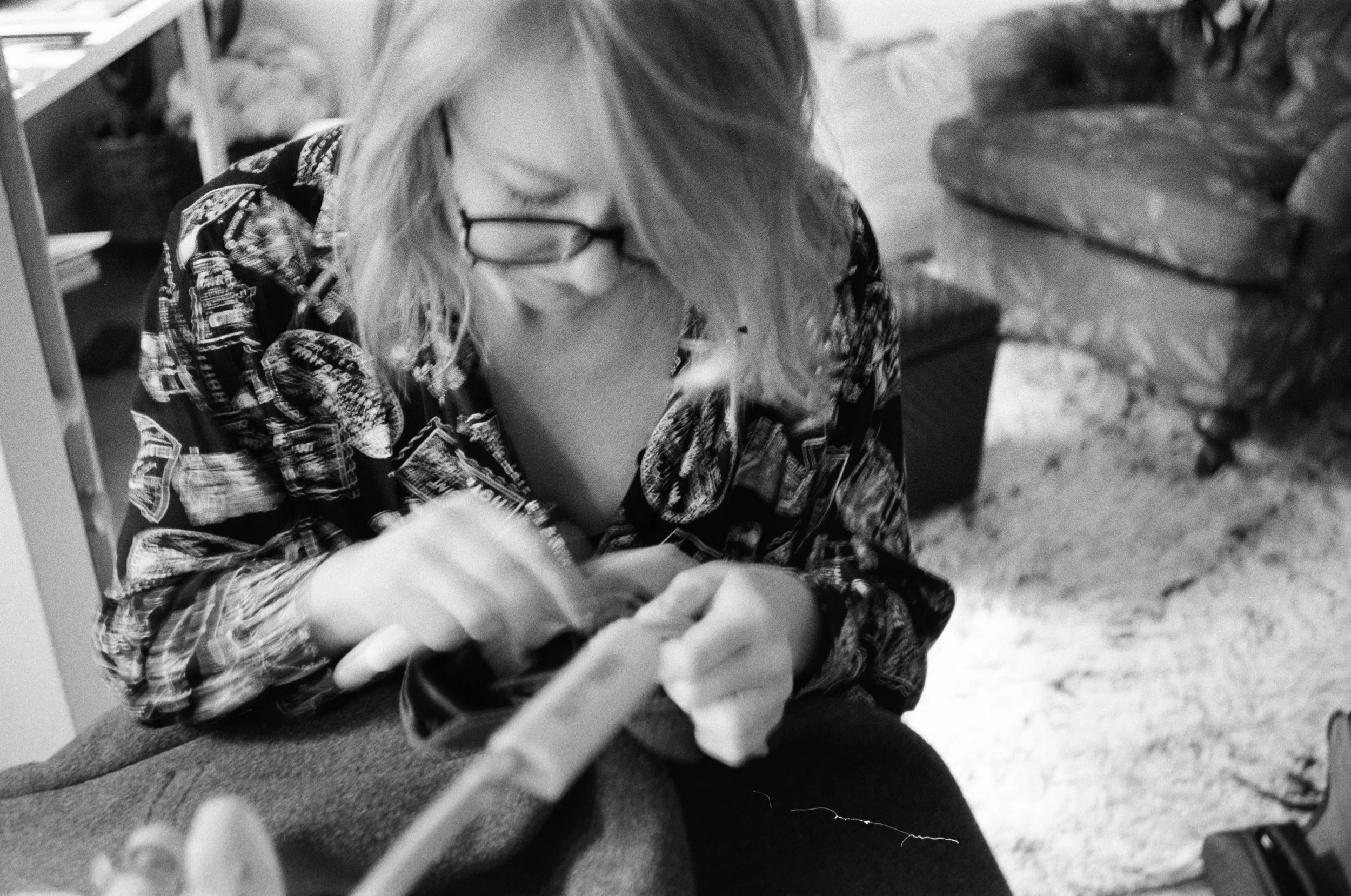 A person with glasses and long hair, wearing a patterned shirt, working on alterations at her sewing machine.