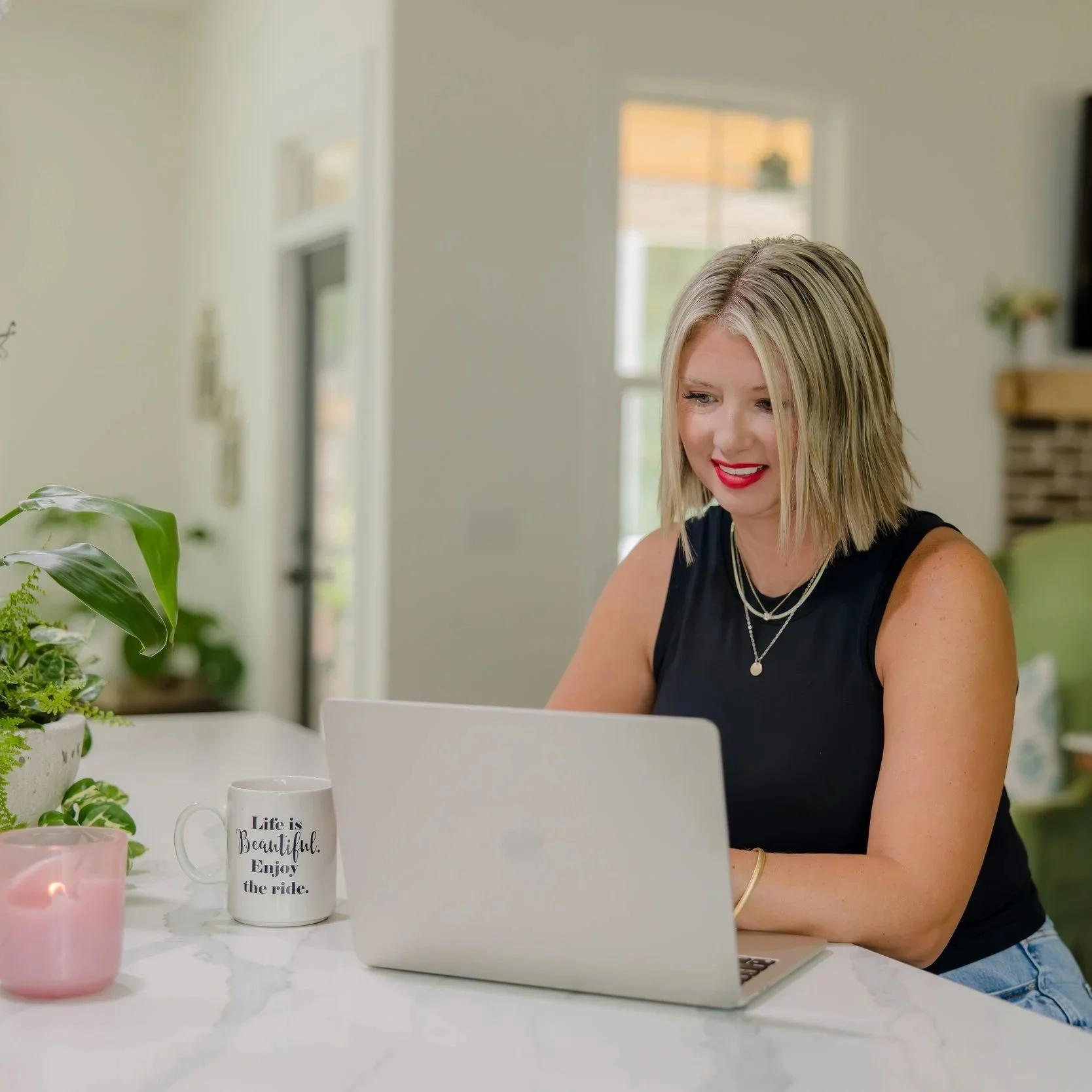 A woman with blonde hair and red lipstick sitting at a white marble table using a laptop in a bright, cozy room with plants and kitchen decor.