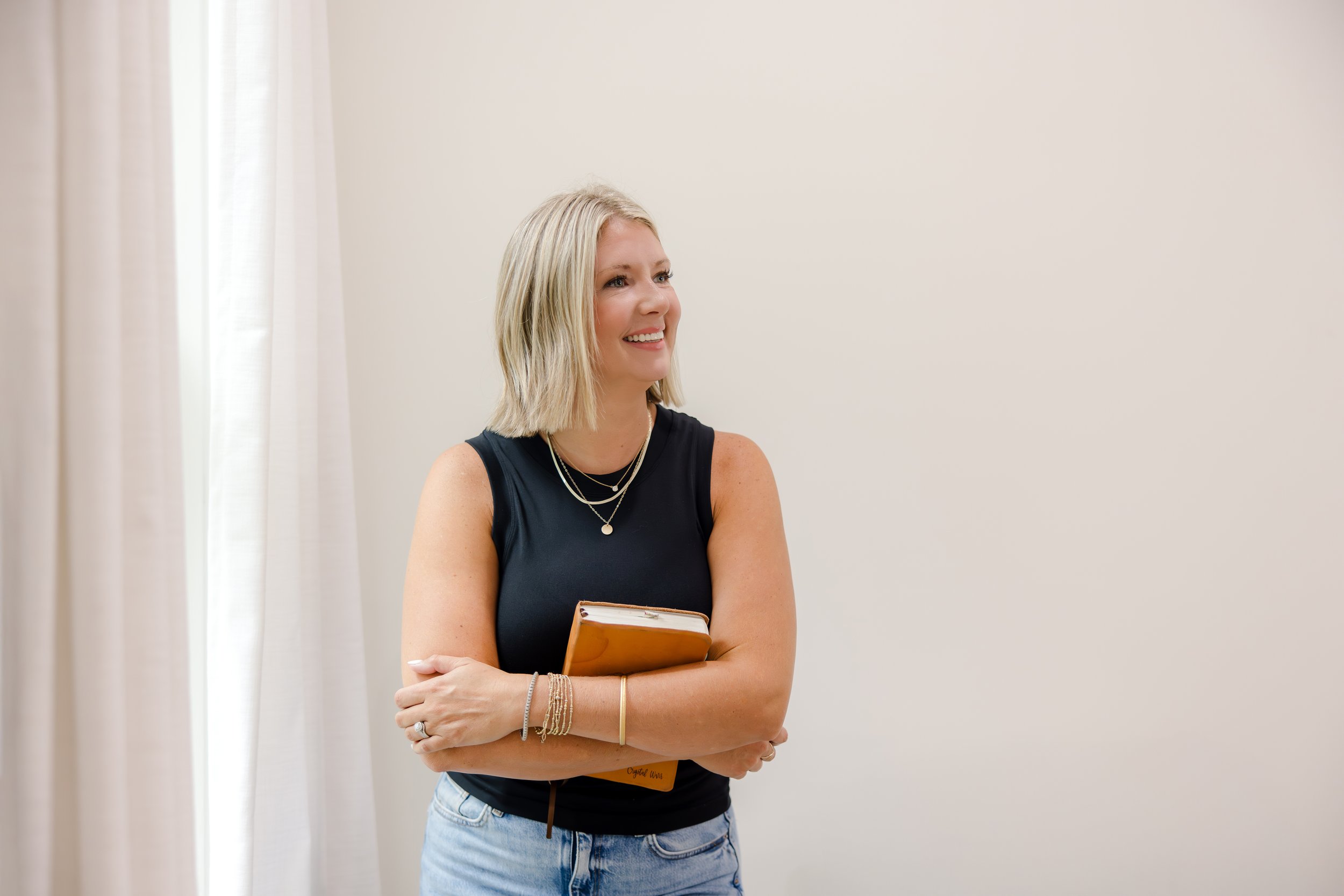 A smiling woman with blonde hair wearing a black sleeveless top and jeans, standing indoors near a cream wall, holding a Bible