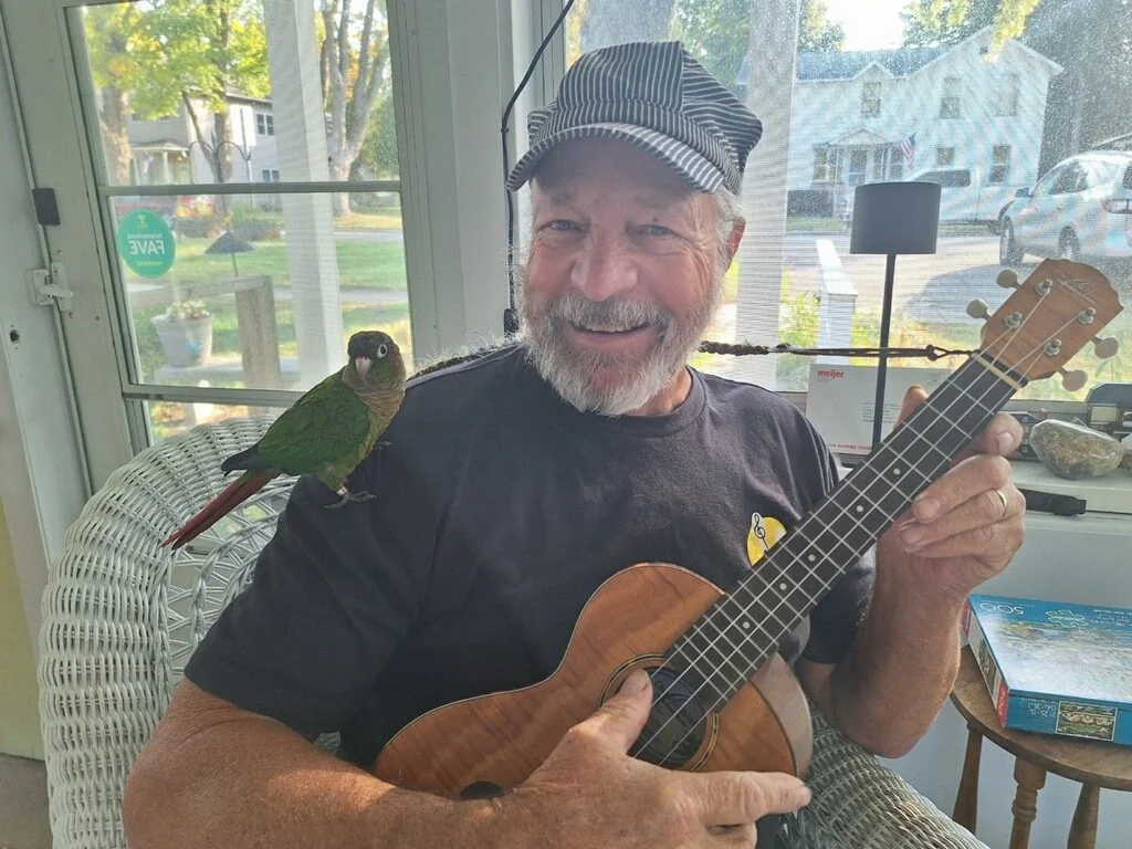 Man sitting on a chair with a small green parrot on his shoulder, holding a ukulele, wearing a striped conductor hat, inside a sunlit room with glass windows.