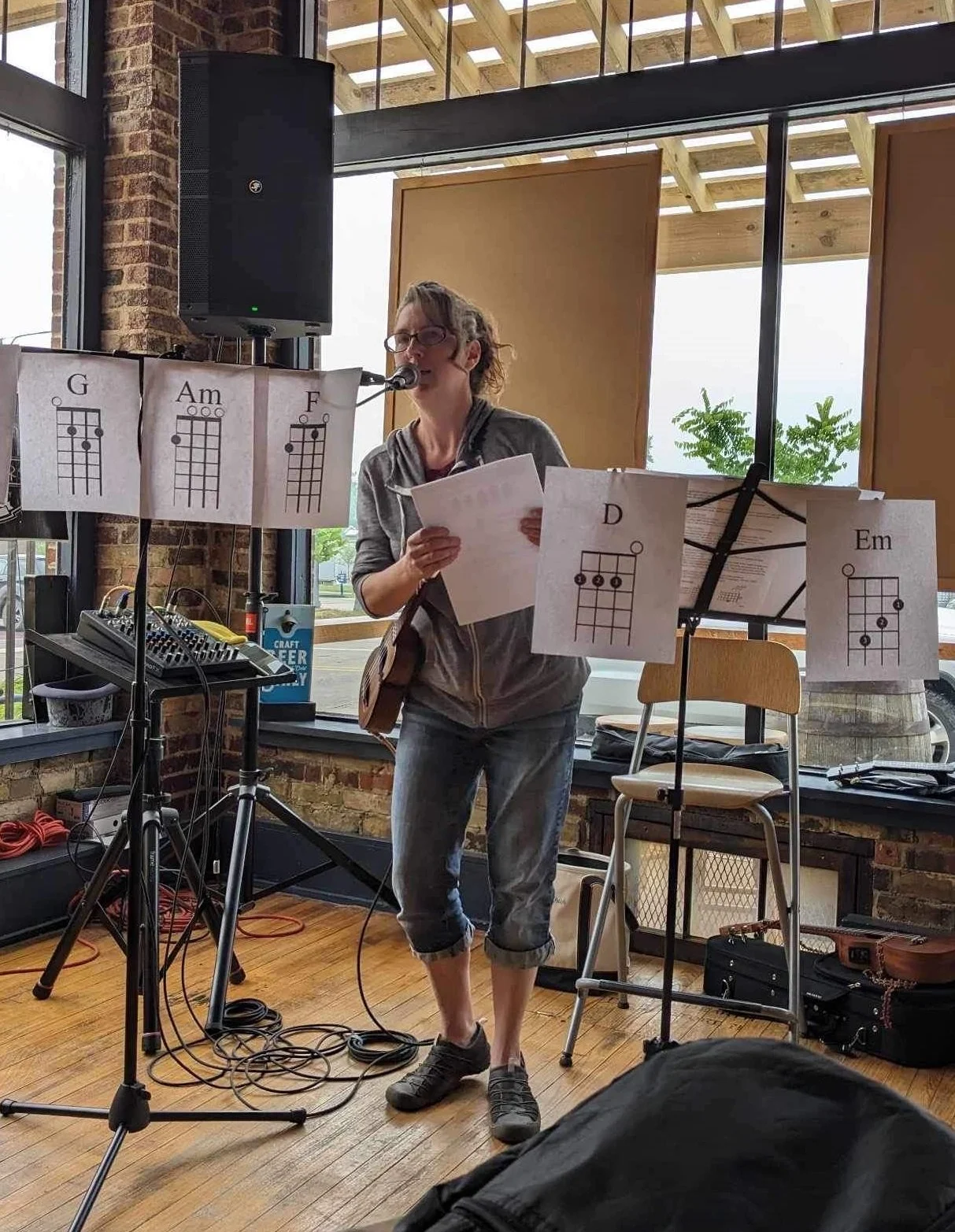 Person standing indoors, playing a ukulele with chord charts displayed. Microphone and audio equipment visible.