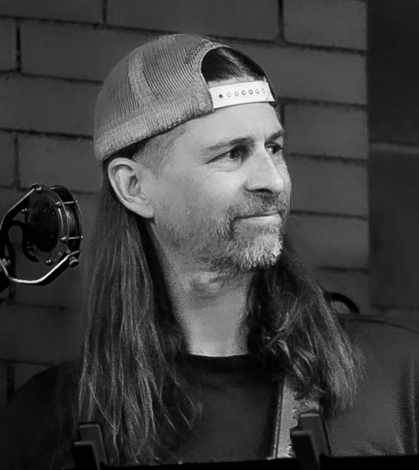 Black and white photo of a person with long hair wearing a backward cap, standing in front of a brick wall with a microphone nearby.