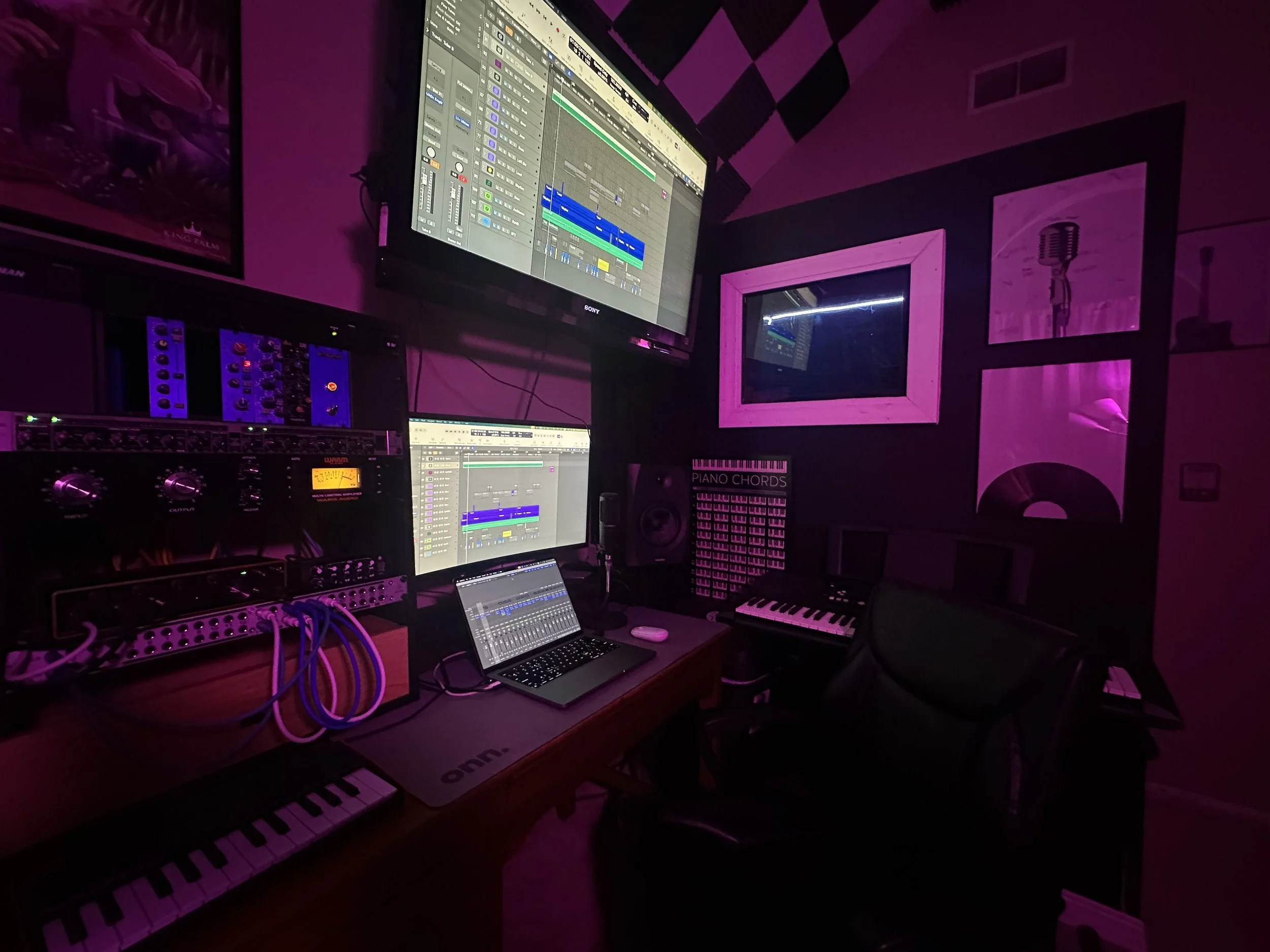 Home recording studio setup with large monitors, a keyboard, and a laptop. Audio mixing software is displayed on screens. The room has purple and orange lighting with soundproofing panels and music equipment.