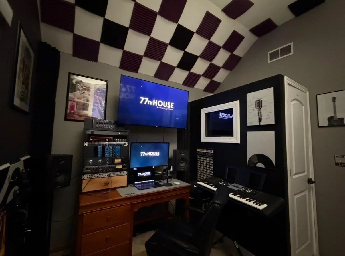 Home recording studio setup with large monitors, a keyboard, and a laptop. Audio mixing software is displayed on screens. The room has purple and orange lighting with soundproofing panels and music equipment.