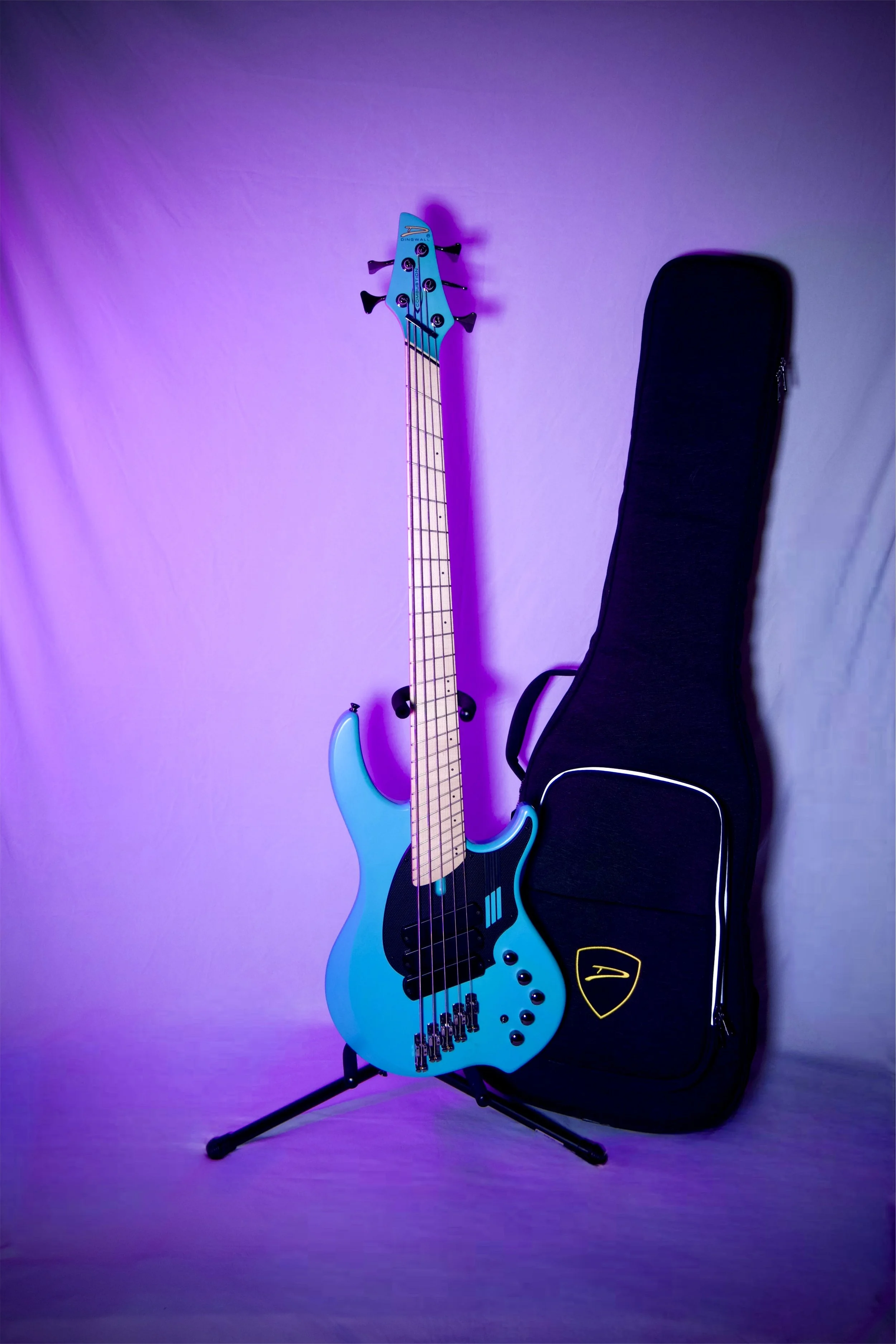 A turquoise electric bass guitar with a natural wood neck and black pickguard, standing on a stand against a quilted white backdrop.