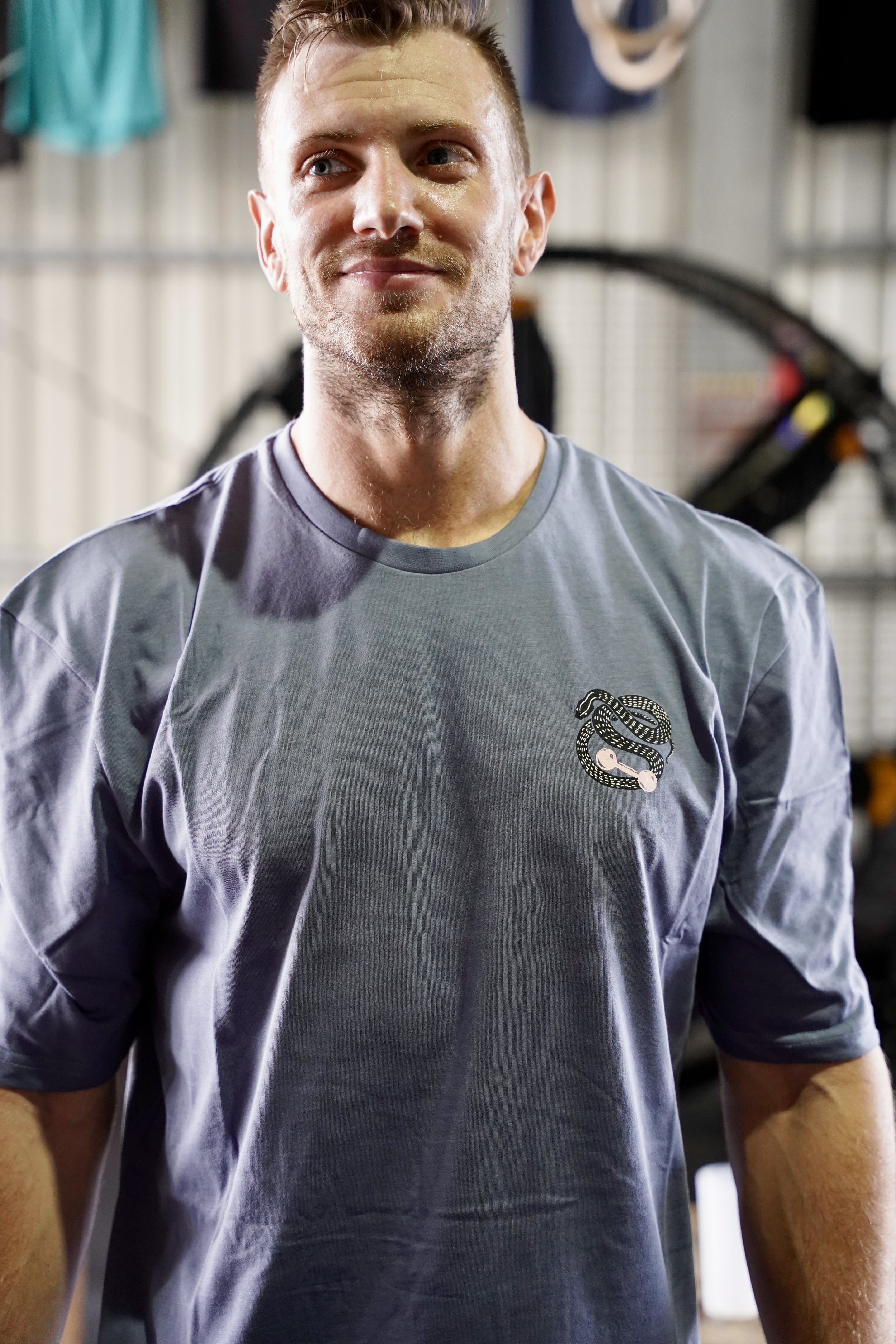 L’image montre un homme souriant et habillé d’un t-shirt bleu avec un logo sur le cœur qui est un serpent