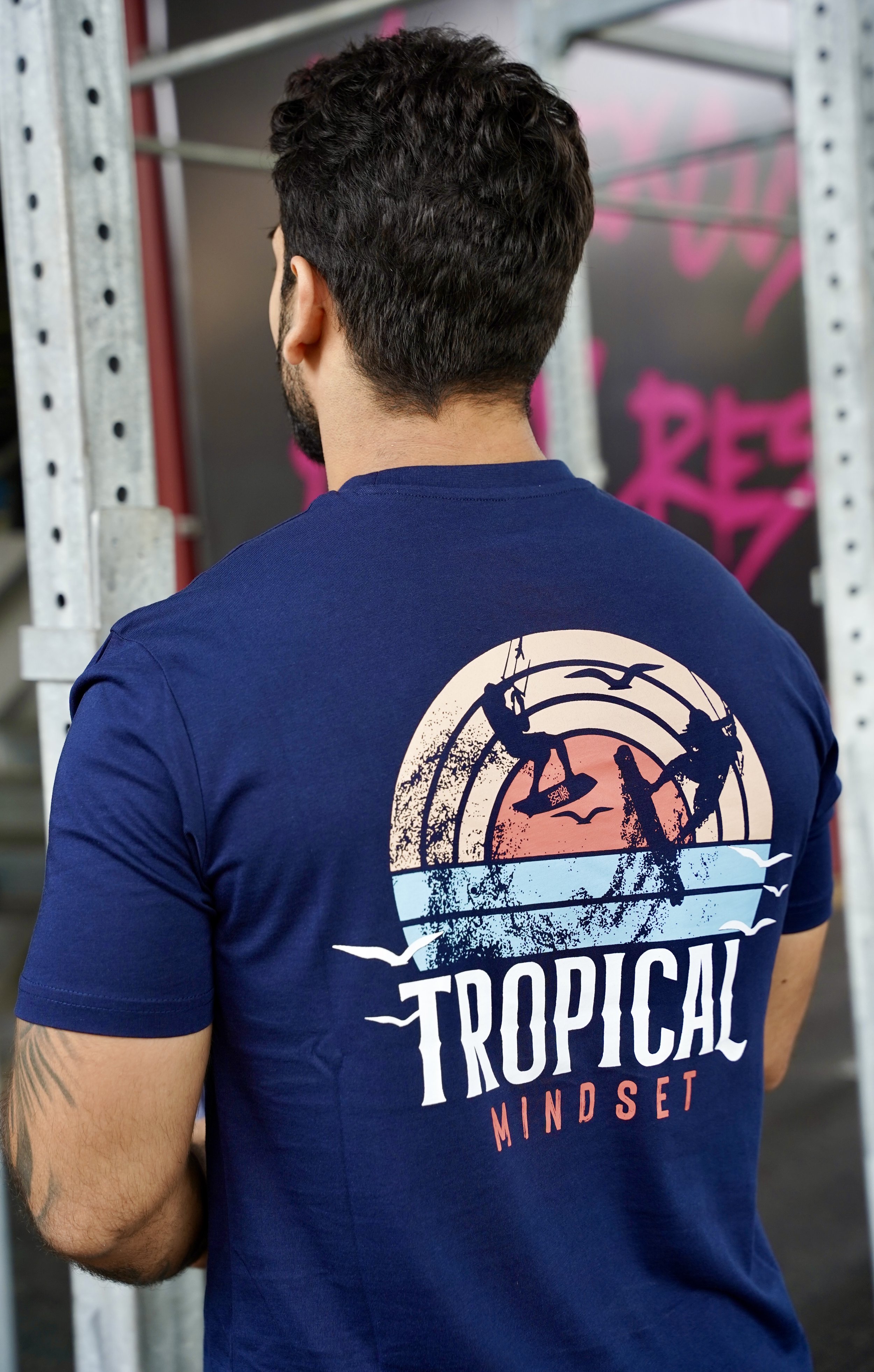 L'image montre un homme de dos portant un t-shirt bleu avec un design coloré. Le motif sur le t-shirt comporte des silhouettes de personnes faisant du kite surf avec le texte "Tropical Mindset".