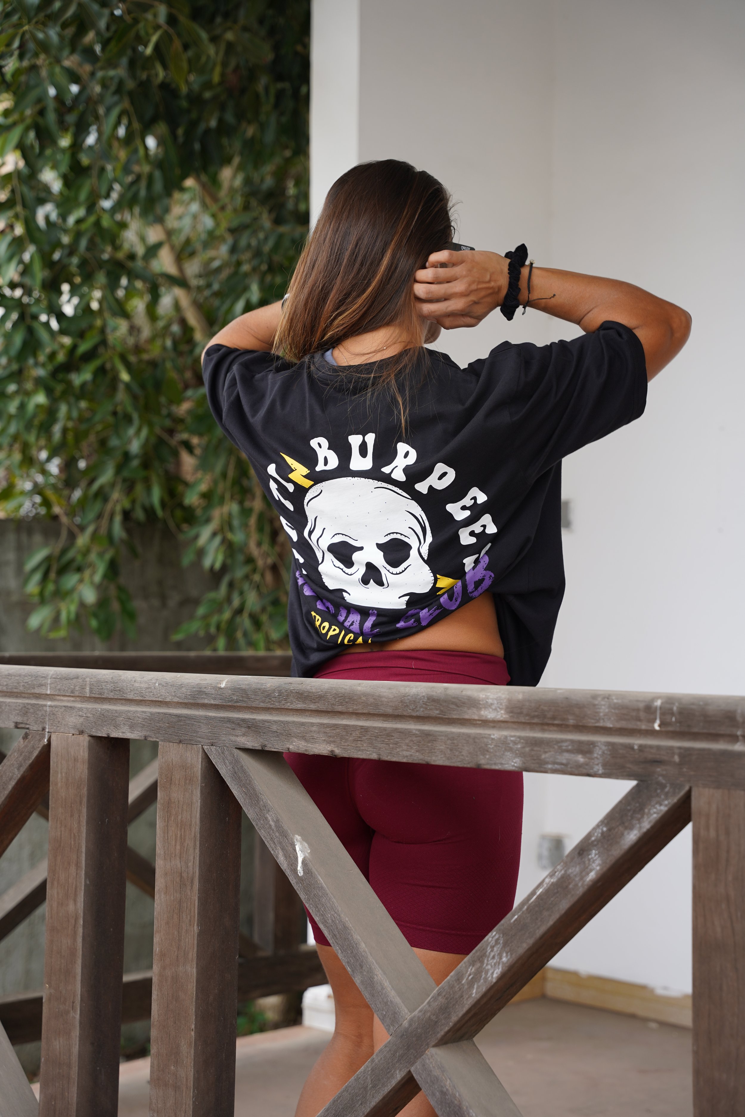 L’image montre une femme de dos assise sur un mur et vêtue t-shirt noir avec une tête de mort oú il est noté anti burpees social club