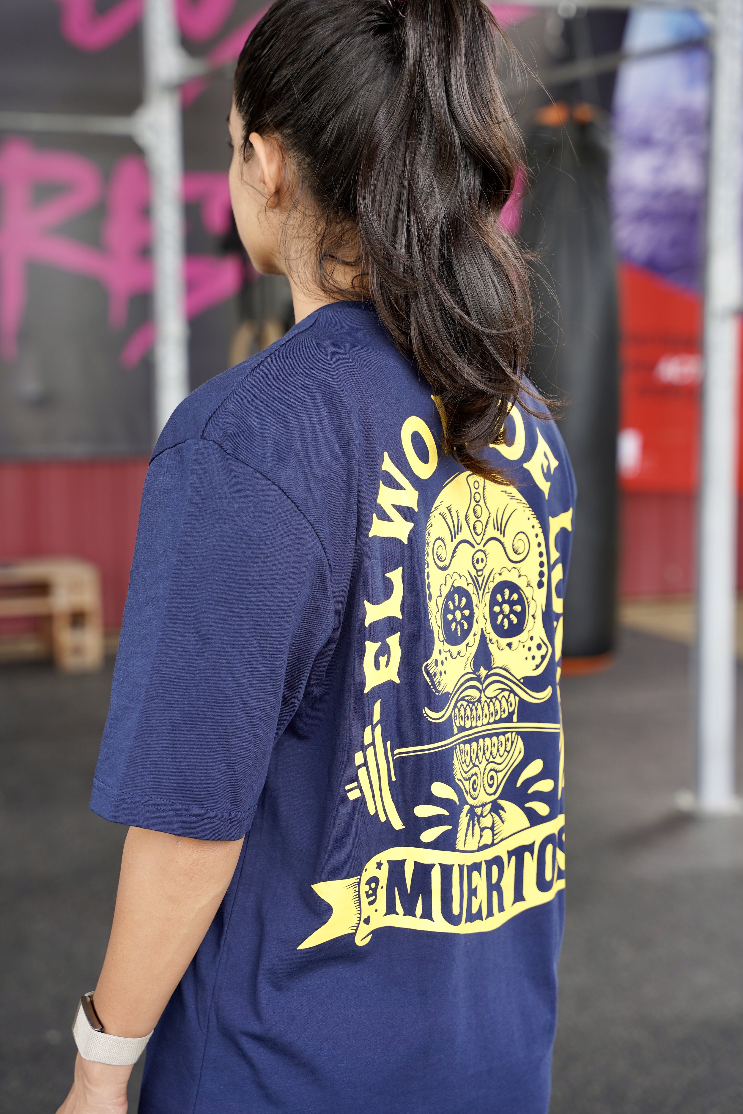 L’image montre une femme vêtue d’un t-shirt bleu avec un logo de tête de mort jaune sur le dos oú il est écrit el wod de los muertos