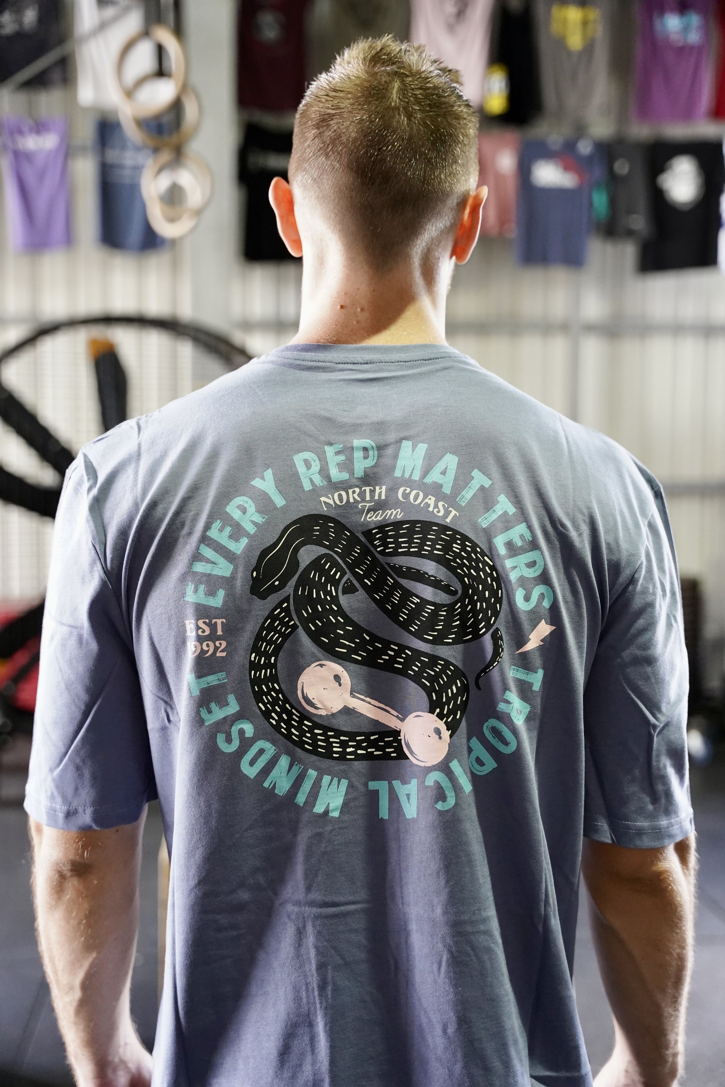 L’image montre un homme de dos et habillé d’un t-shirt bleu qui représente un serpent et un haltère