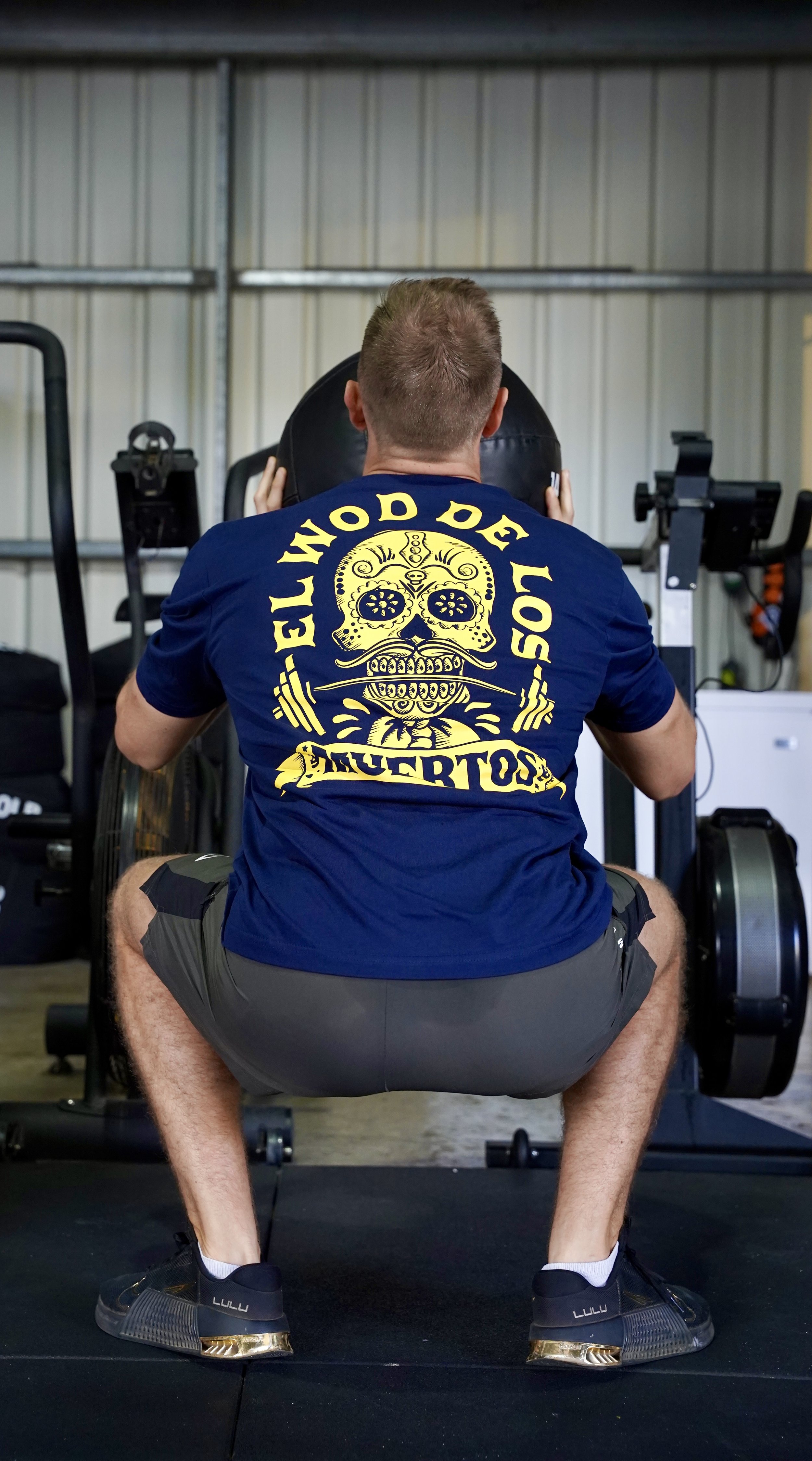 L’image montre un homme en position squat faisait des exercices et il est habillé d’un t-shirt bleu foncé avec un logo de tête de mort