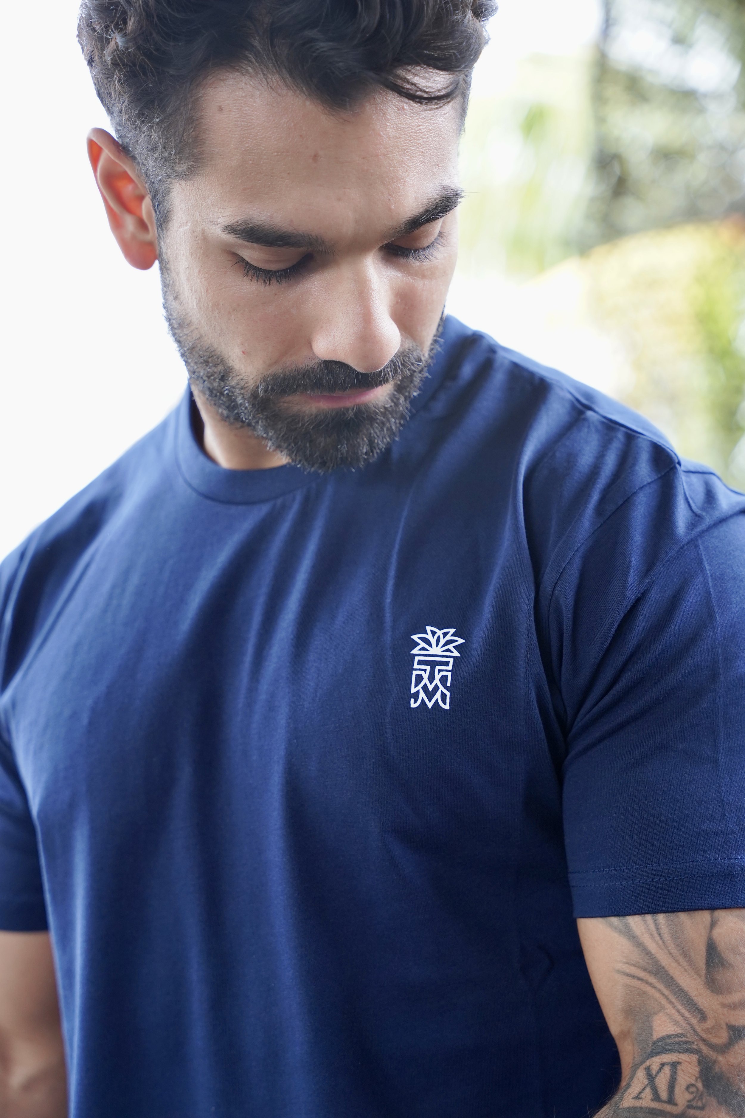 L'image montre un homme portant un t-shirt bleu avec un petit design de palmier sur la poitrine. Le fond est flou, mettant en valeur le t-shirt.