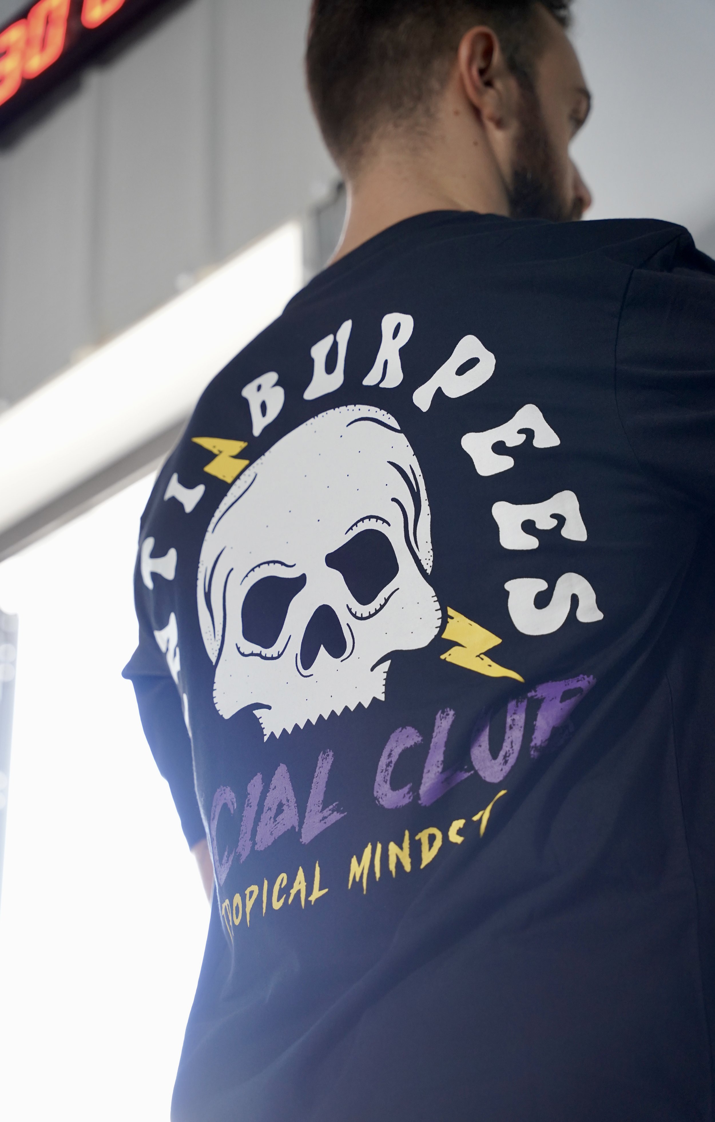 L’image montre un homme de dos vêtu t-shirt avec une tête de mort oú il est noté anti burpees social club