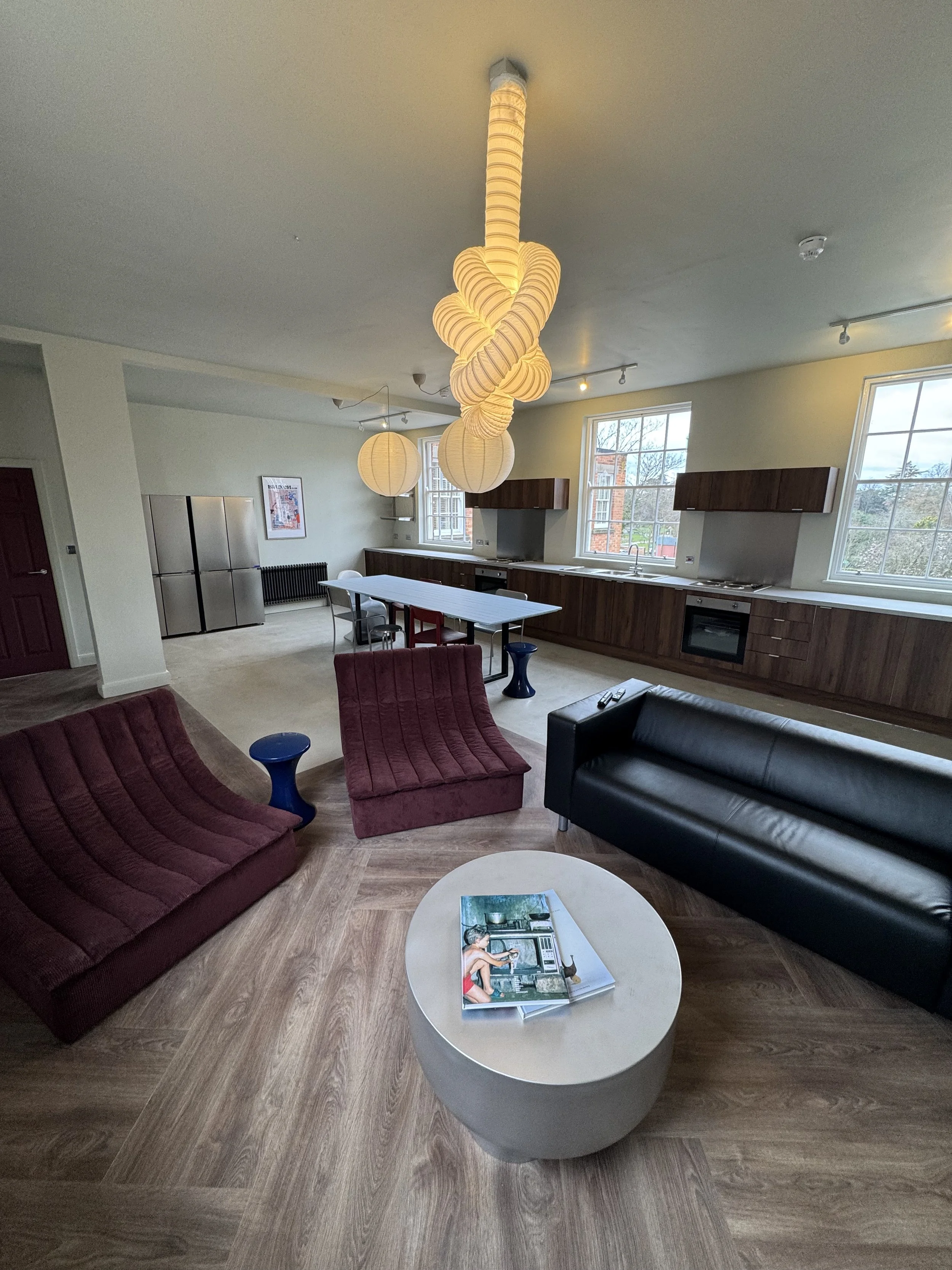 Modern living room with maroon and black sofas, a coffee table with magazines, a dining table, kitchen area with wooden cabinets, stainless steel refrigerator, large windows, hanging lights including a spiral paper lantern, and wooden flooring.