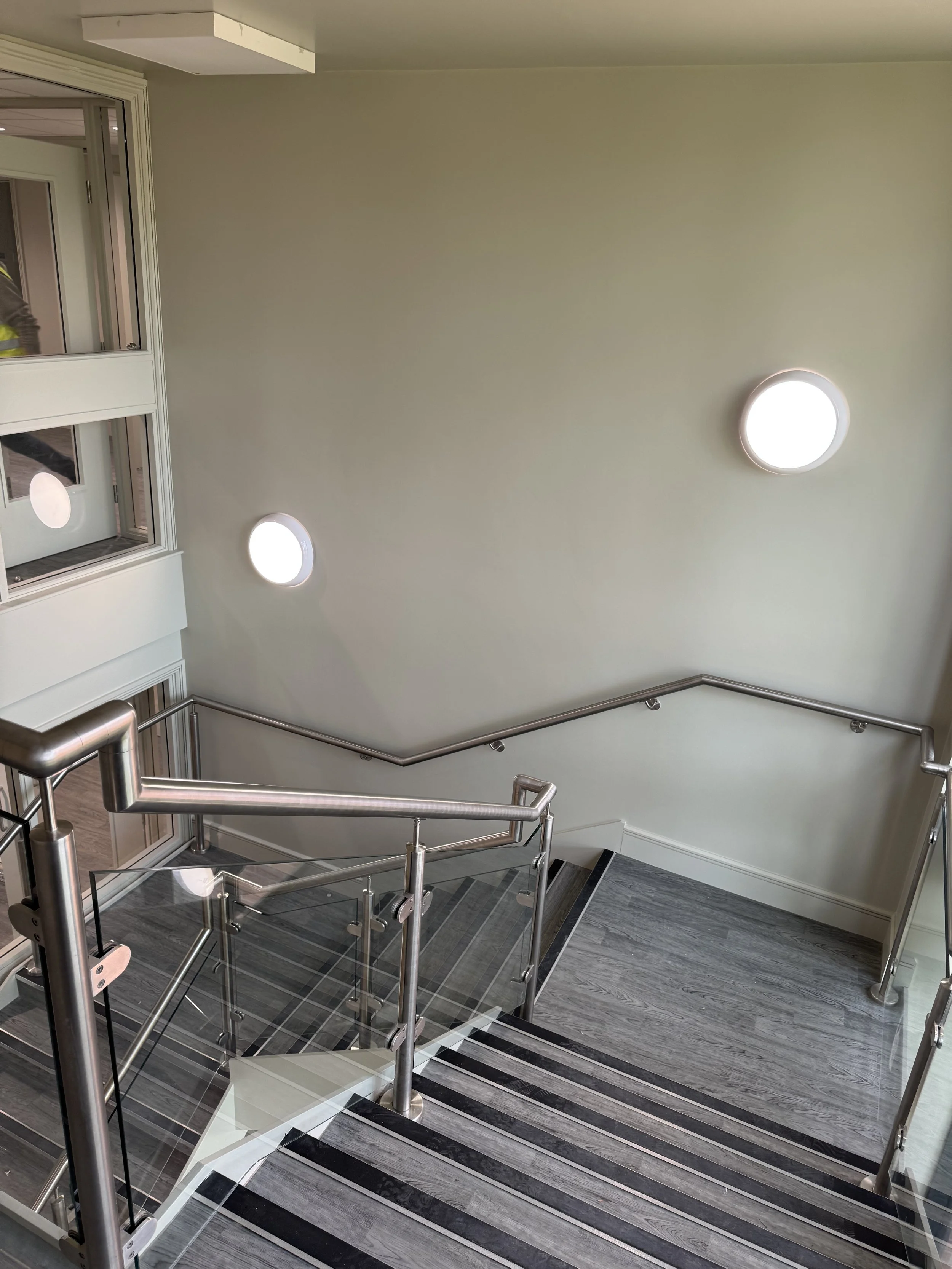 Indoor staircase with stainless steel handrails and glass panels, gray wooden flooring, white walls with two circular wall lights, and a window showing part of an exterior balcony.