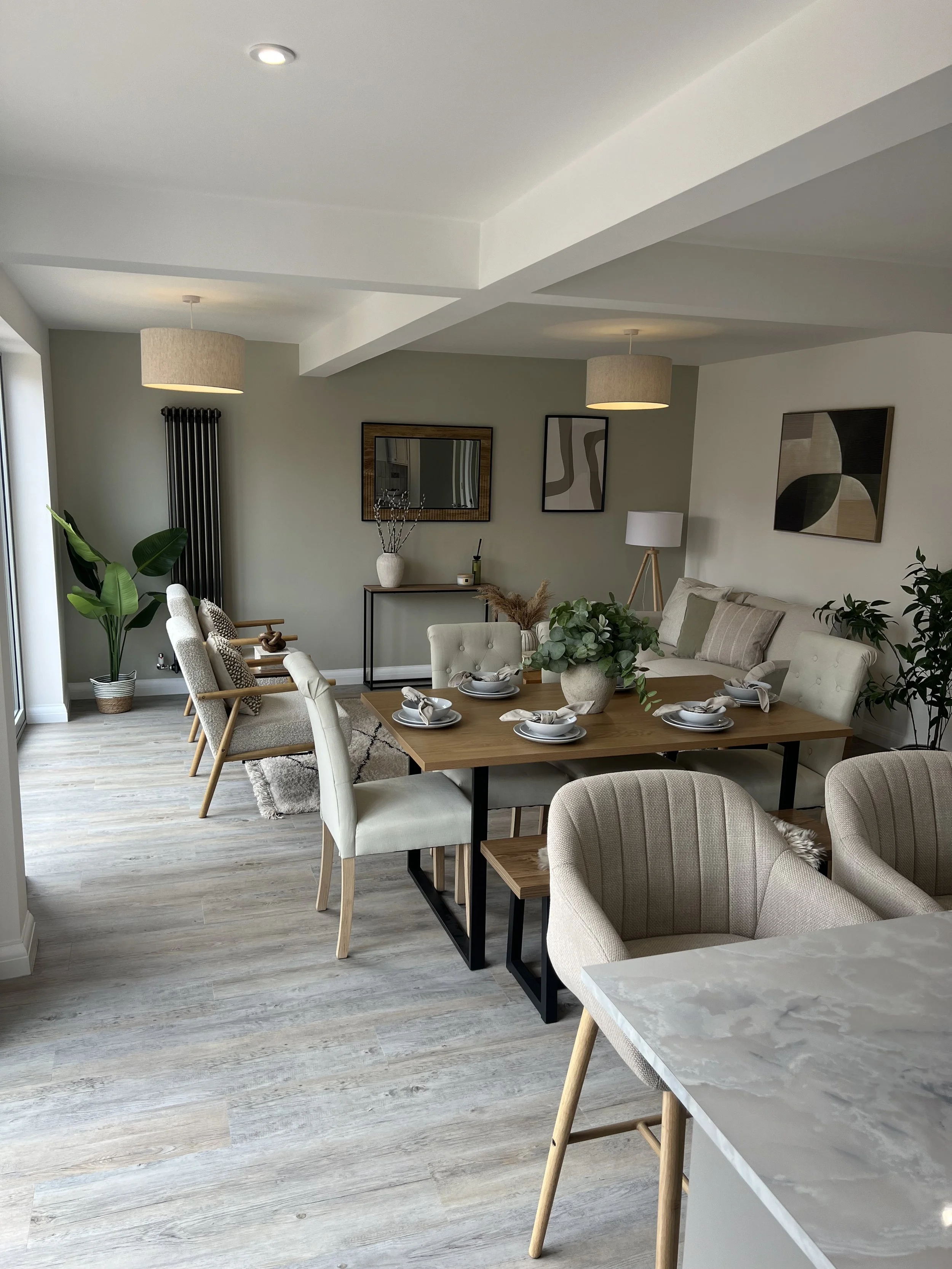 Modern living room and dining area with light-colored furniture, wooden accents, wall art, large potted plants, and a marble kitchen counter.