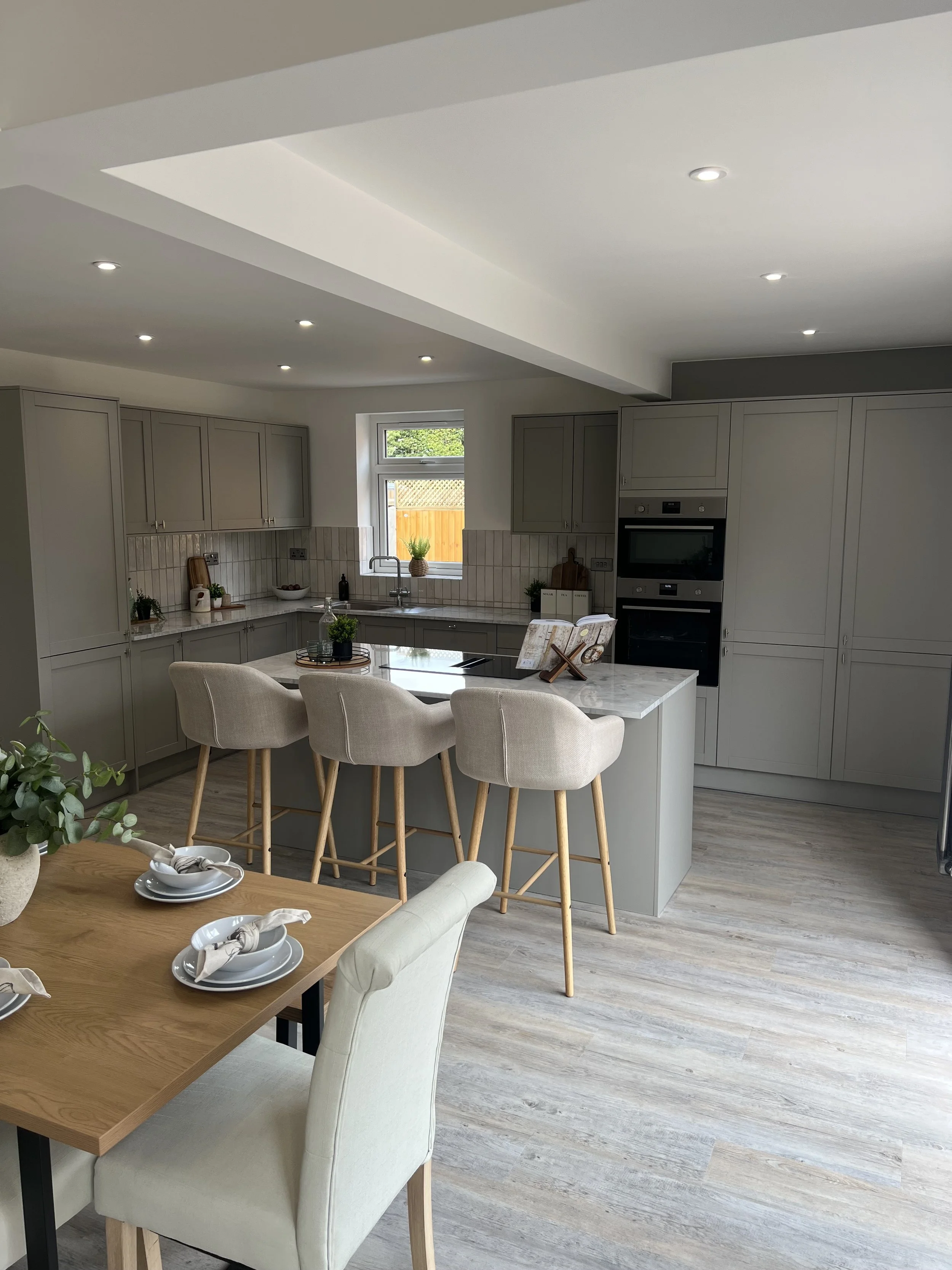 A modern kitchen with gray cabinets, a marble island, and a dining table set with white plates, napkins, and a centerpiece, featuring a window with greenery outside and recessed ceiling lights.