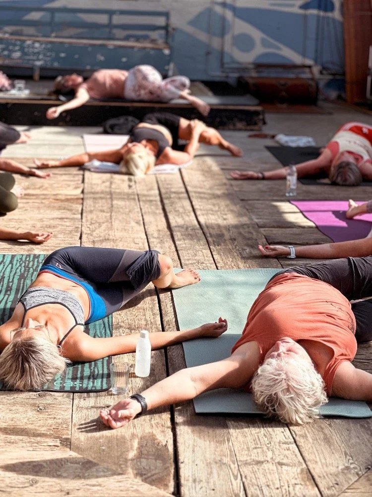 Människor som utövar yoga eller meditation på en träfacad, liggande på yogamattor utomhus i solljus.