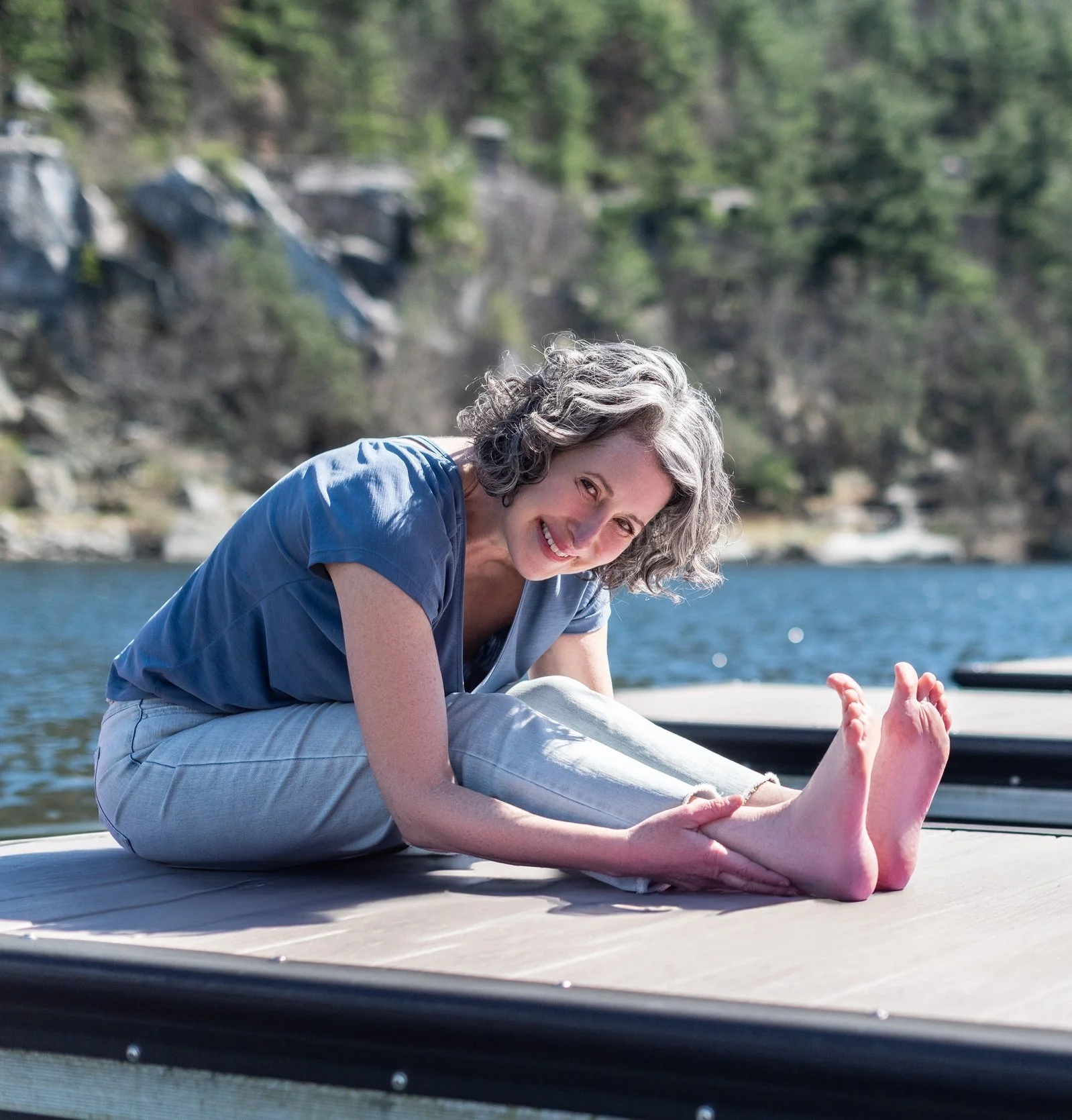 A woman in a blue shirt sitting on a dock by the water, stretching her legs and smiling at the camera.
