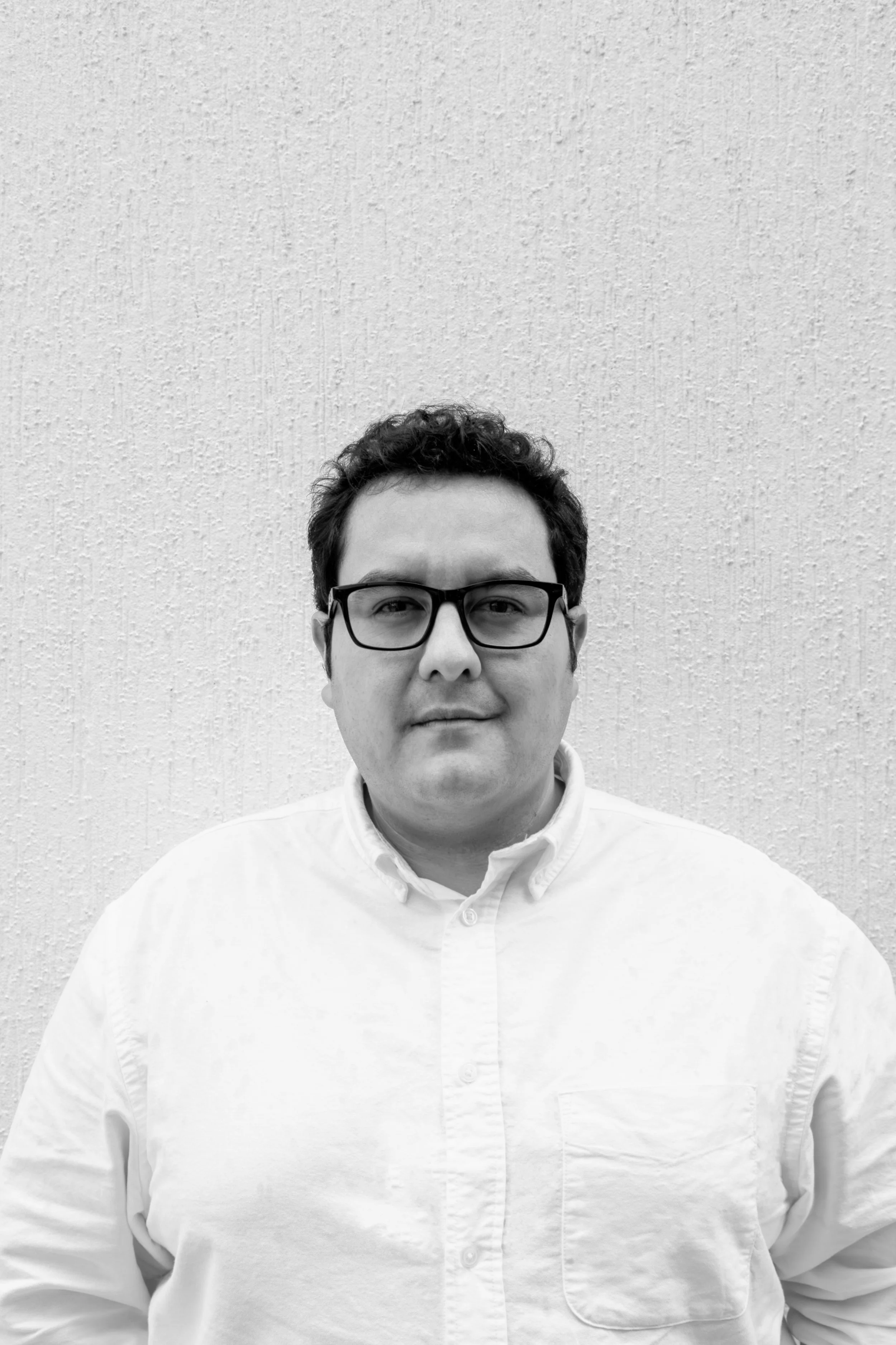 Alejandro Terán Ávalos. Retrato en blanco y negro de un hombre con gafas y camisa blanca, de cabello castaño oscuro, frente a una pared de textura rugosa.  BIOMA arquitectura + paisaje. Arquitecto, Estudio de Arquitectura, Quito, Ecuador.