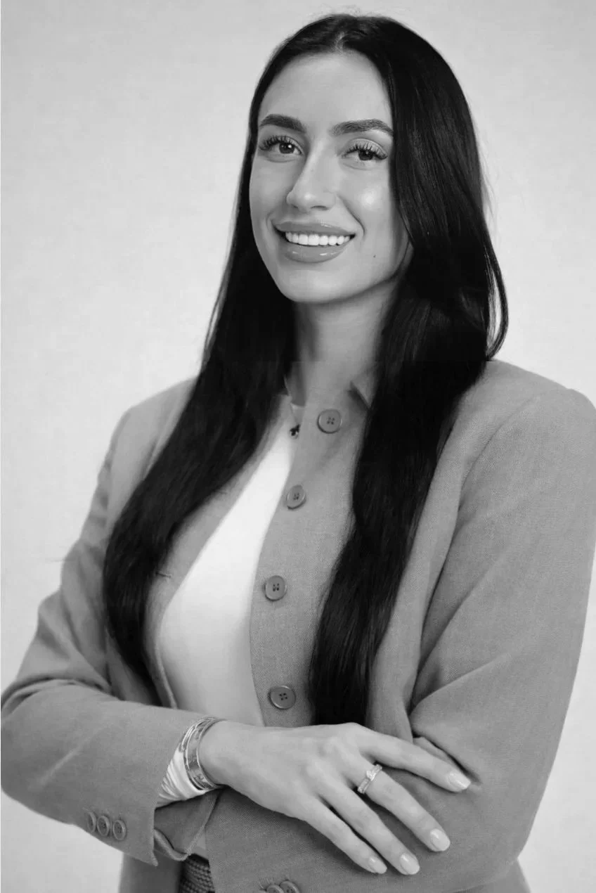 Black and white portrait of a woman with long dark hair, smiling, wearing a blazer with large buttons over a light top, standing with arms crossed.