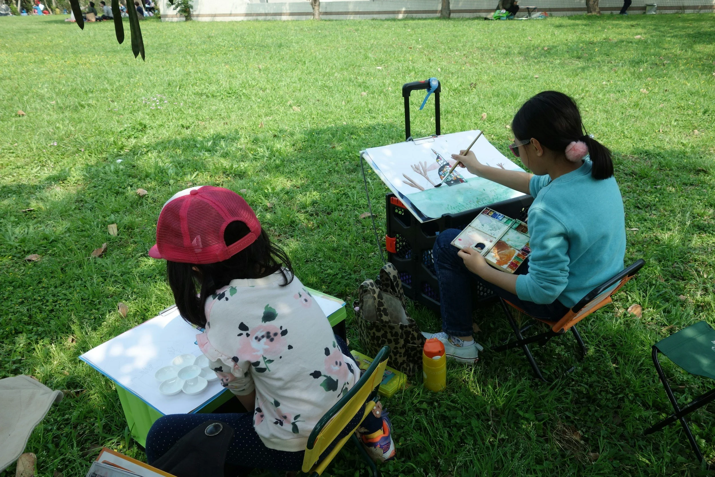 Two children seated on the grass, painting and drawing outdoors. One girl has a pink cap and floral shirt, and the other girl has glasses and pink fluffy earmuffs, painting a scene with a tower and trees.