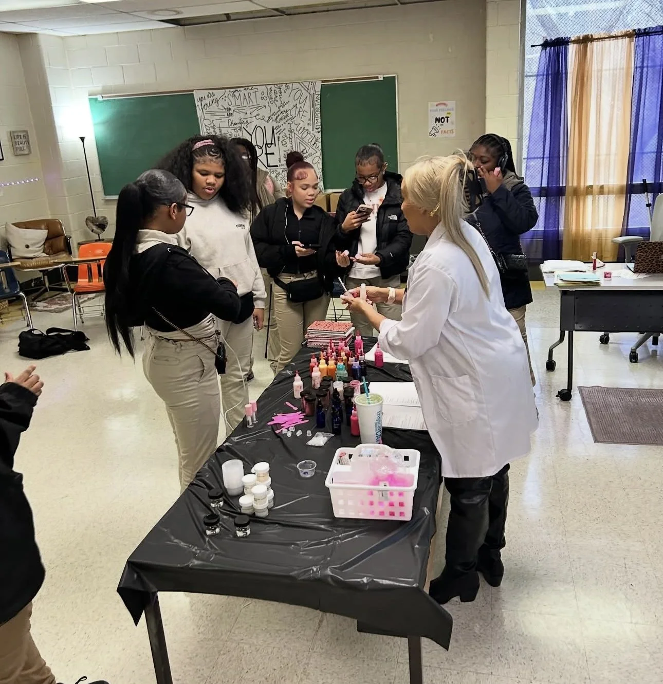 Group of women gathered around a table with nail polish bottles, receiving a demonstration from a woman in a white lab coat in a classroom setting.