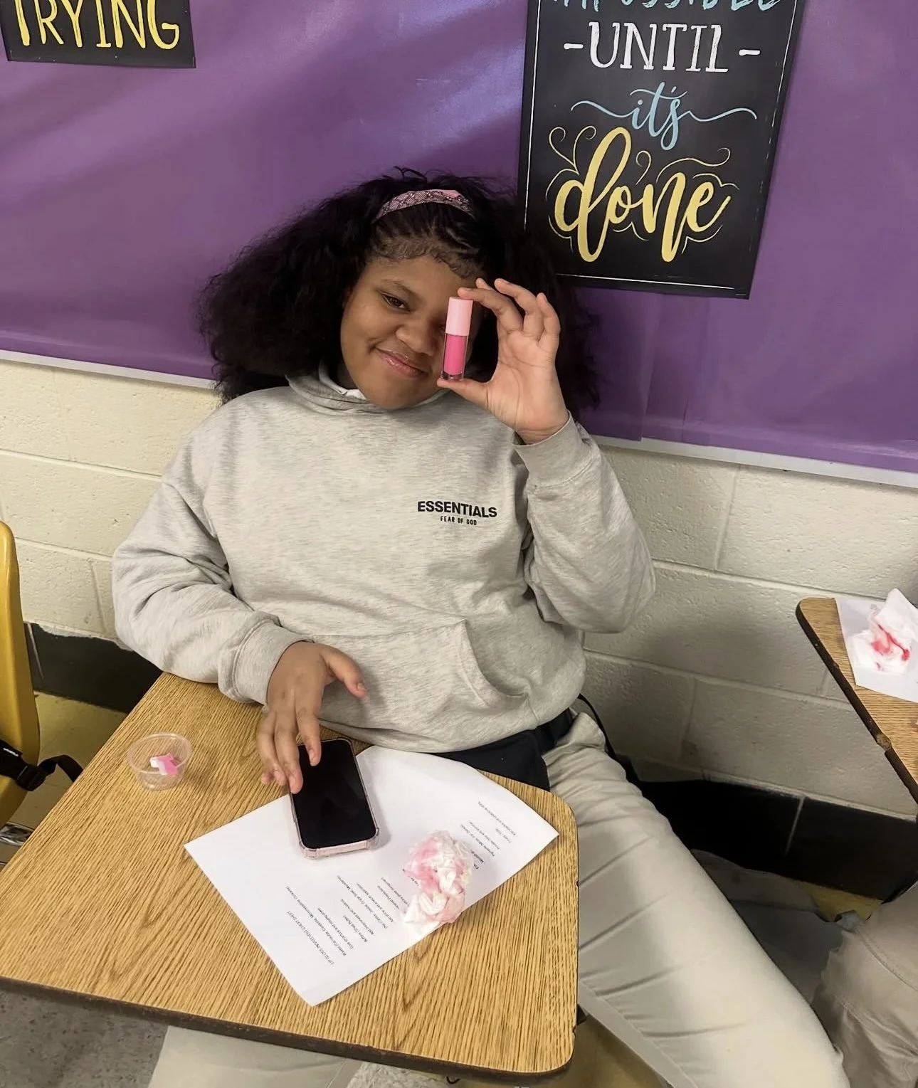 A girl sitting at a desk holding a tube of pink lipstick close to her face while smiling at the camera. There are papers and a phone on the desk, with torn tissue paper nearby. The background has a purple bulletin board with a sign that reads 'Until it's done'.