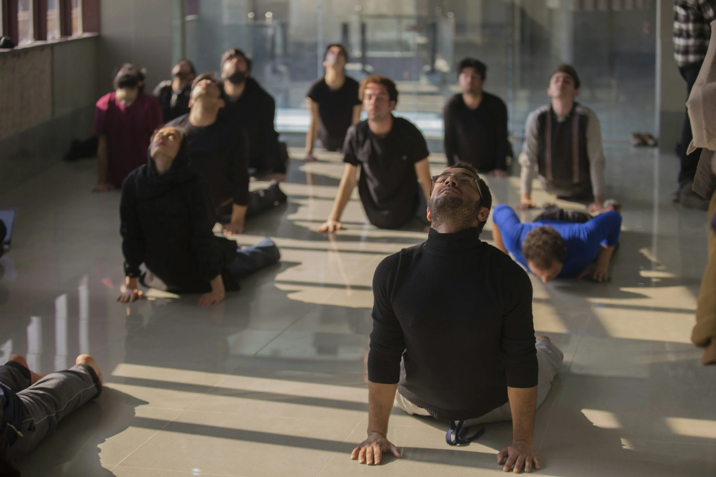 People practicing yoga or stretching indoors in a well-lit space with large windows, sitting or kneeling on the floor.